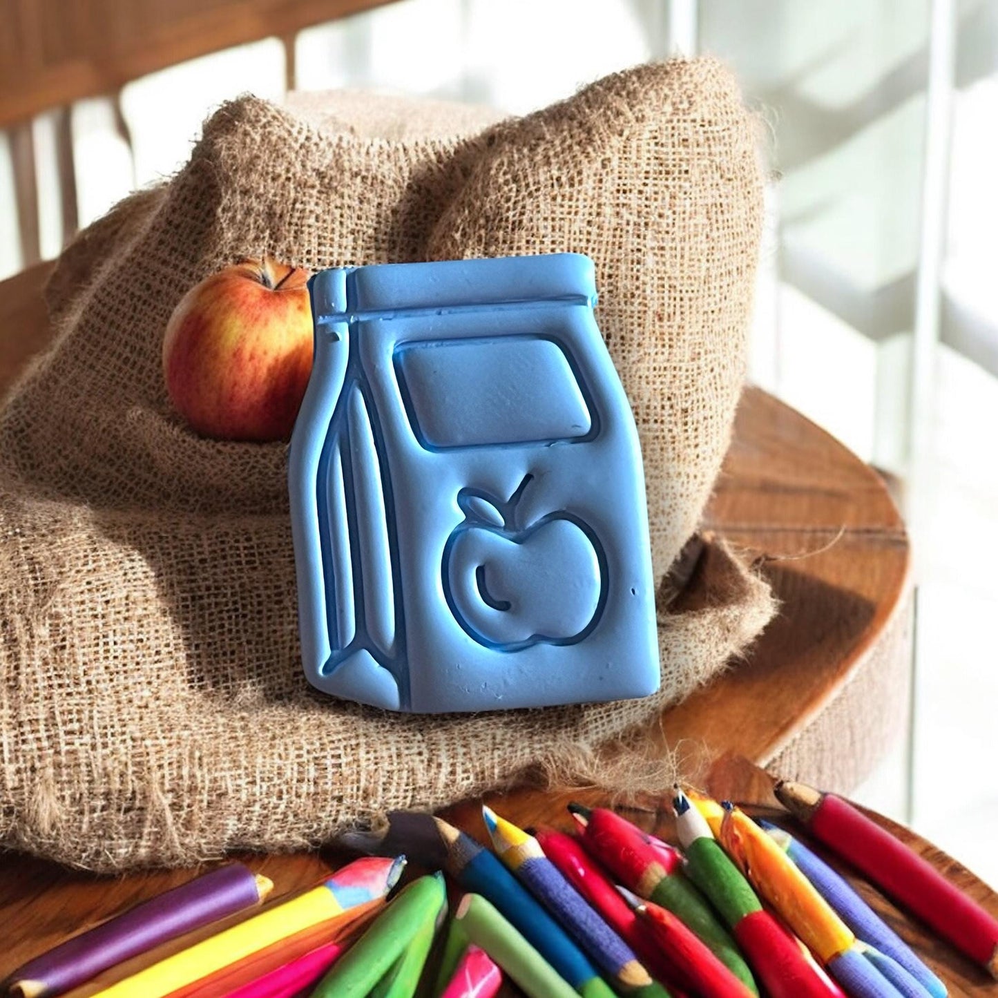 angled clay impression of lunch bag with apple placed near burlap and colored pencils