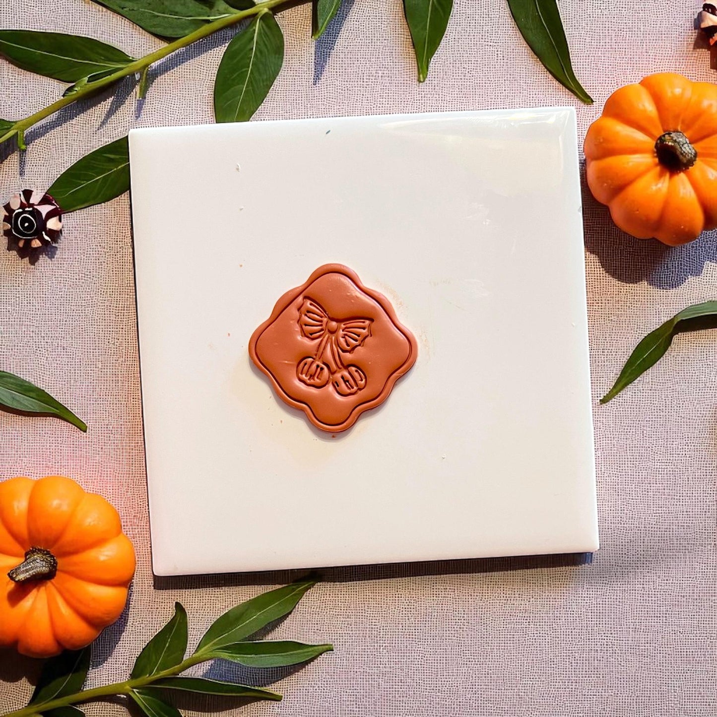 Pumpkin cherry clay cutout centered on a square tile with pumpkins and greenery