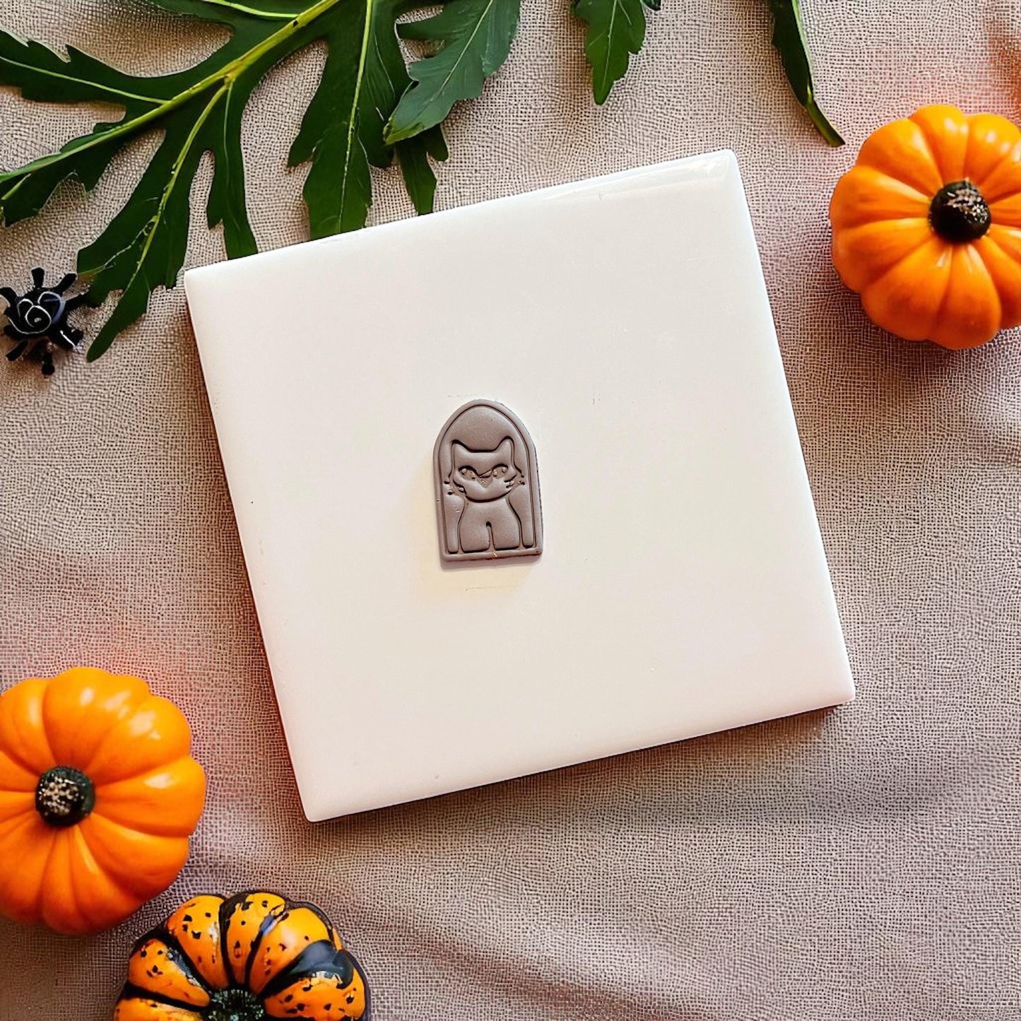 Small clay cutout of spooky cat portrait on white tile surrounded by pumpkins