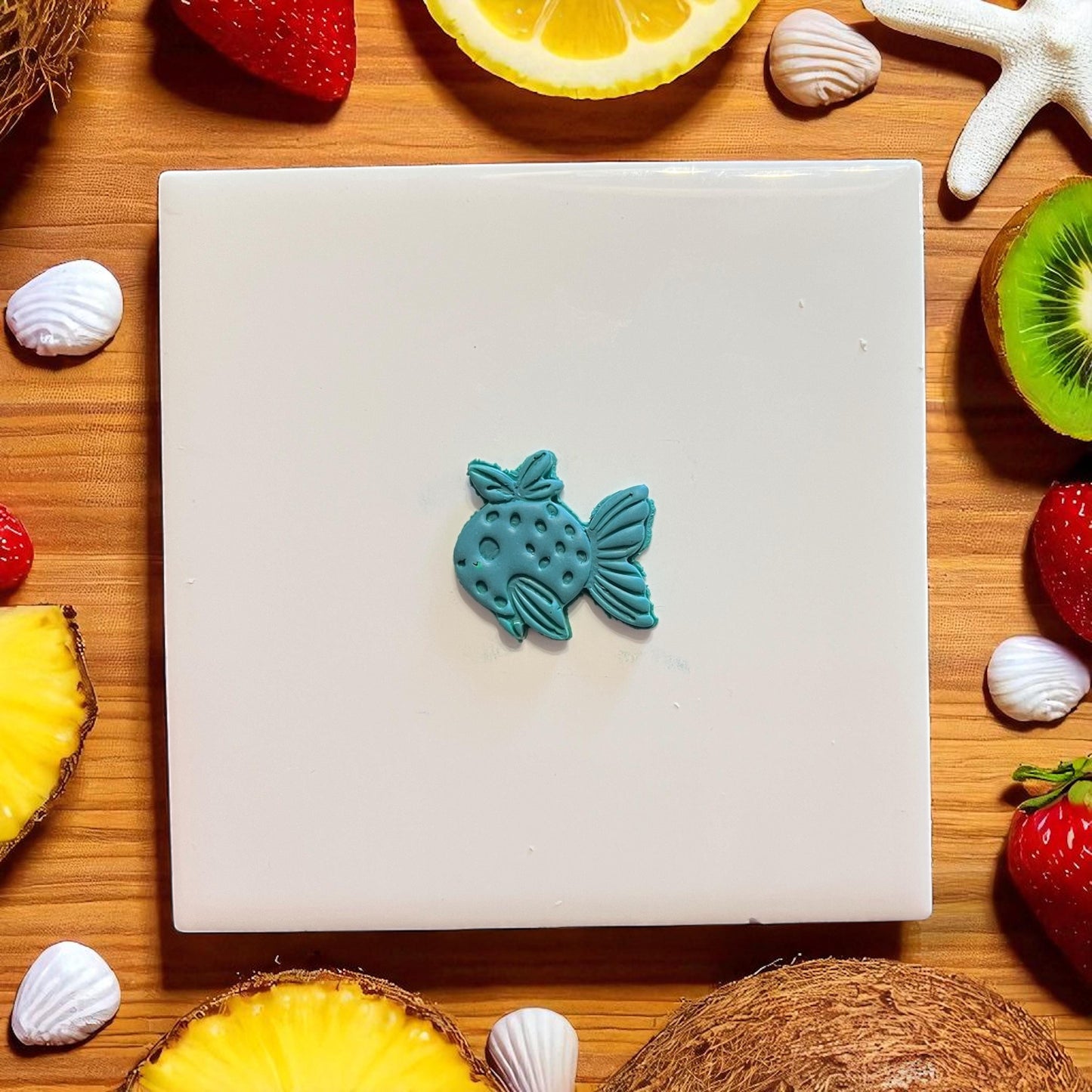Mini strawberry fish shape made with teal clay on tile with fruit background