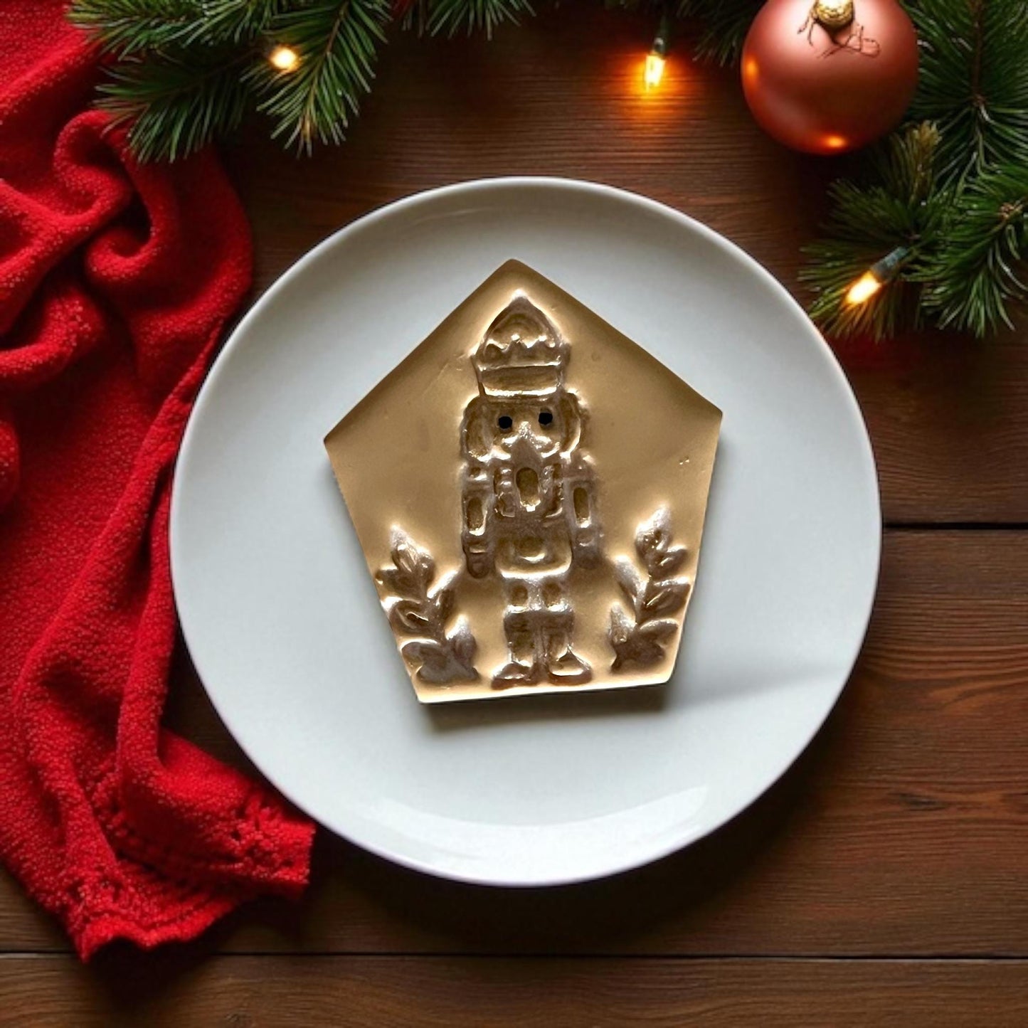 Golden clay imprint of nutcracker soldier with crown and branches on white plate, red fabric, and pine greenery.