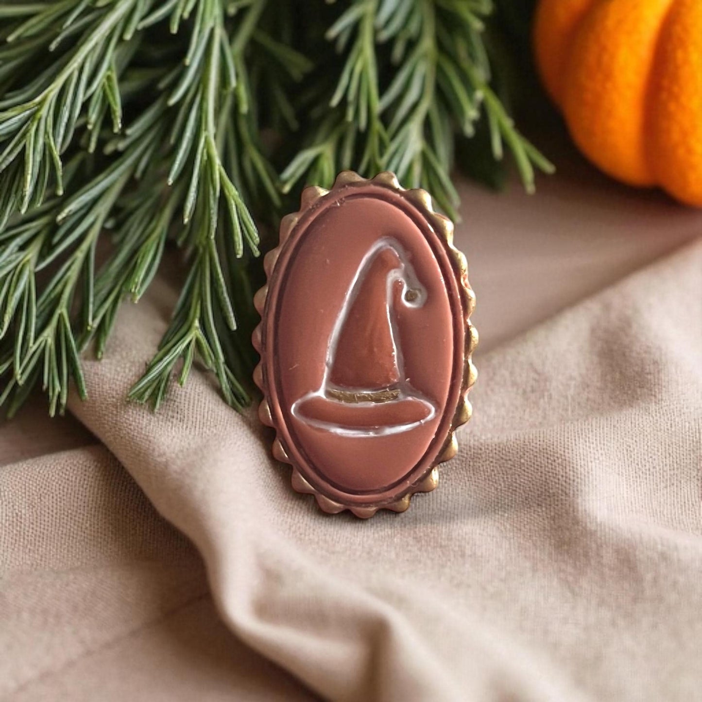 Brown witch hat cameo clay piece with scalloped oval border on fabric with green pine branches.