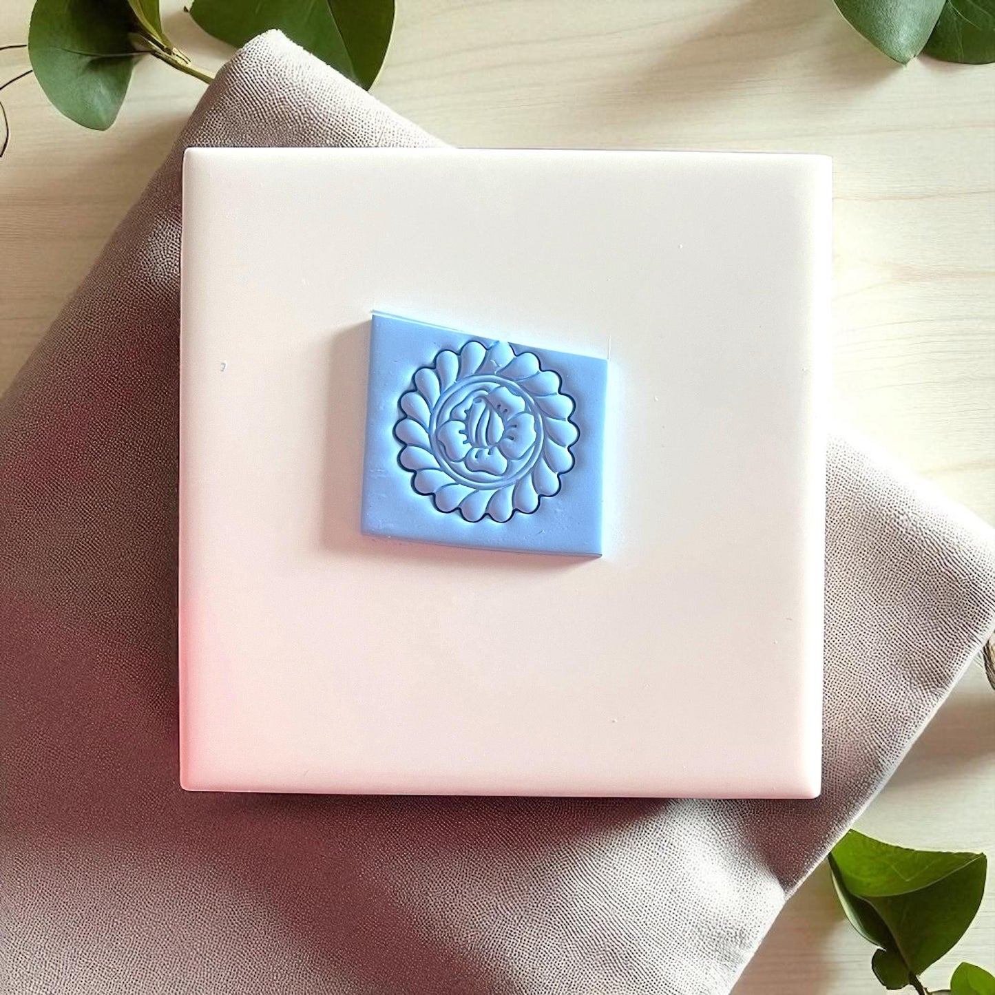 Blue clay imprint of round flower design on white tile with beige fabric and leaves.