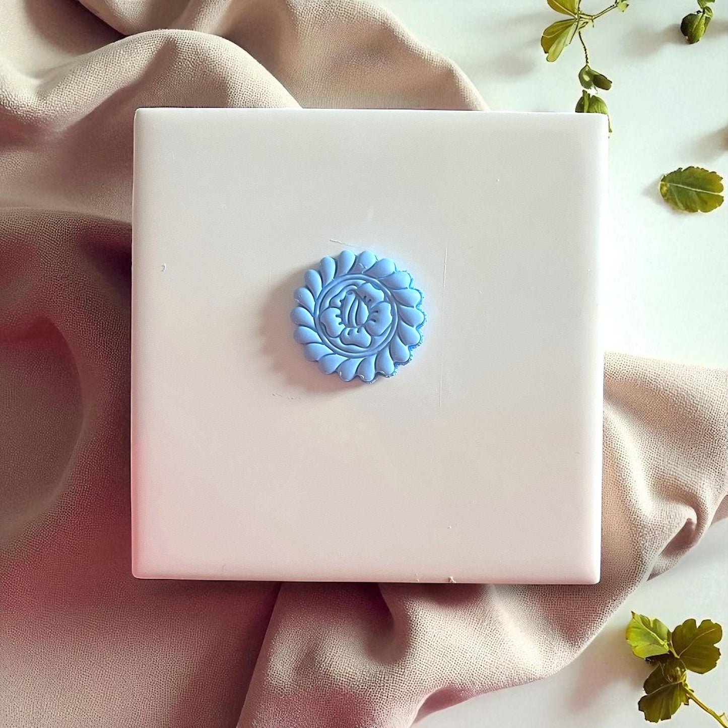 Round blue flower clay imprint centered on white tile with soft fabric and greenery.