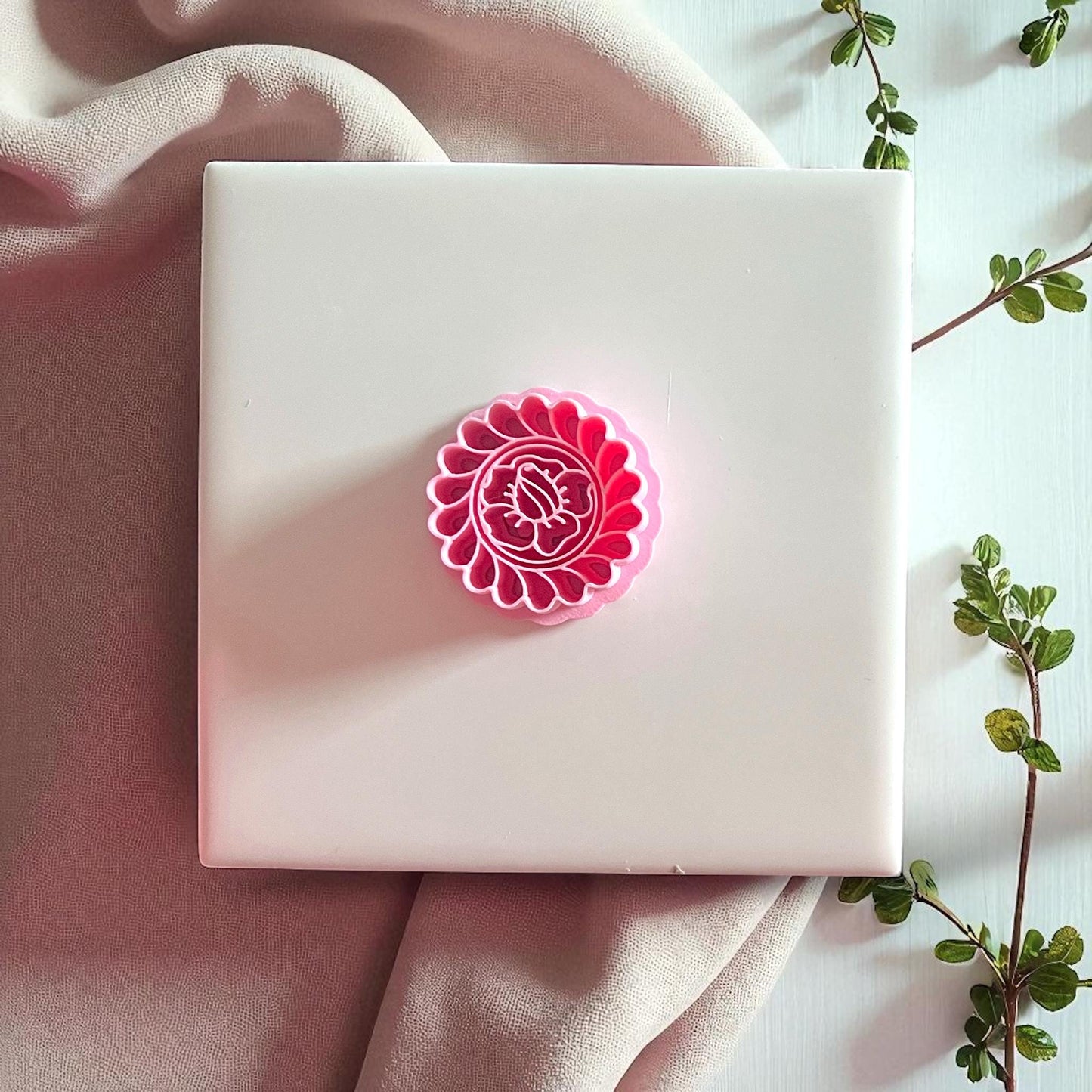 Pink round flower cutter displayed on white tile with beige fabric and greenery.