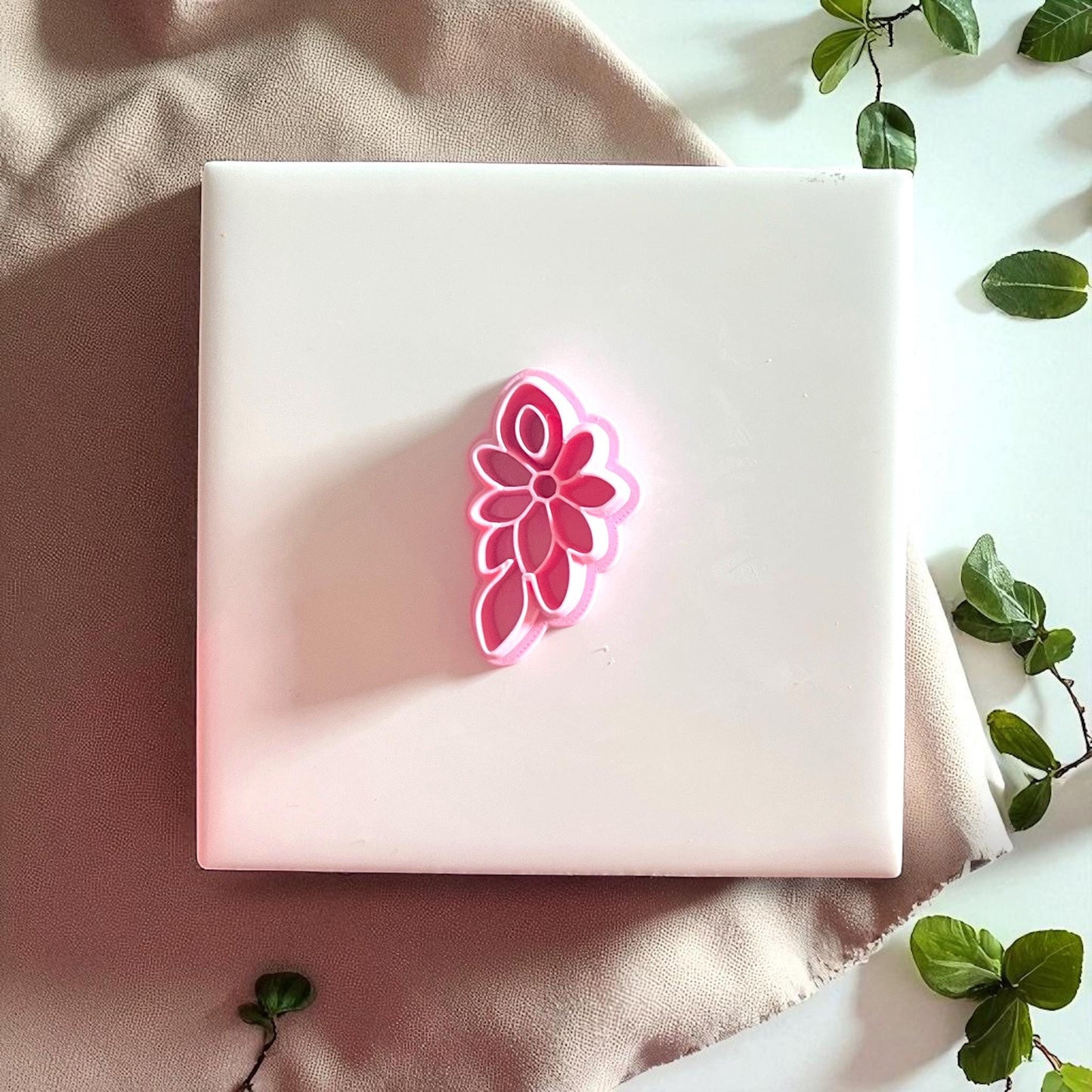 Pink floral cluster cutter displayed on white tile with greenery and fabric.