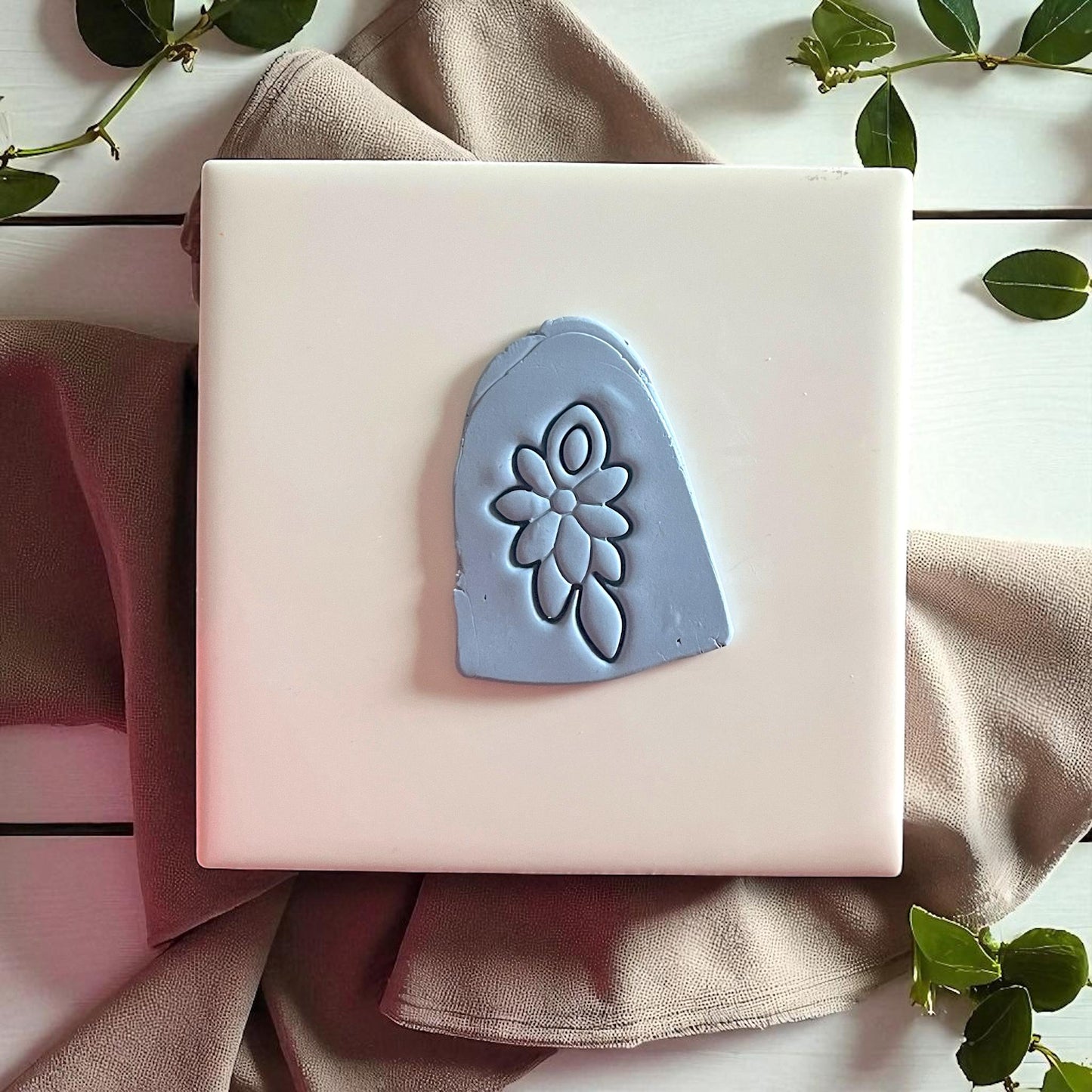 Blue clay imprint of floral cluster design on white tile with fabric and greenery.