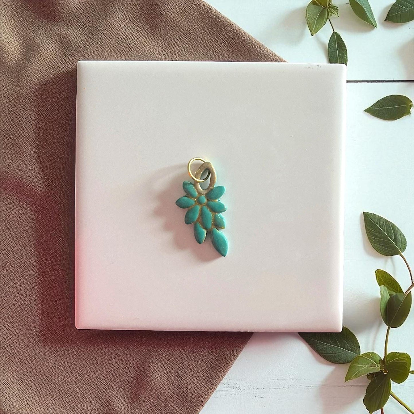 Teal floral clay piece with gold accents and metal loop on white tile with greenery.