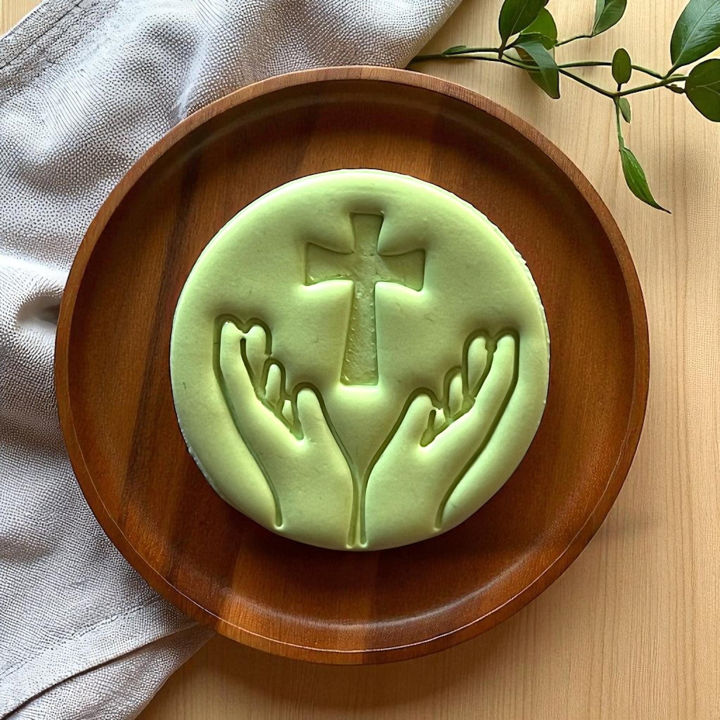 Large green clay imprint of hands holding cross inside wood dish with fabric and greenery.