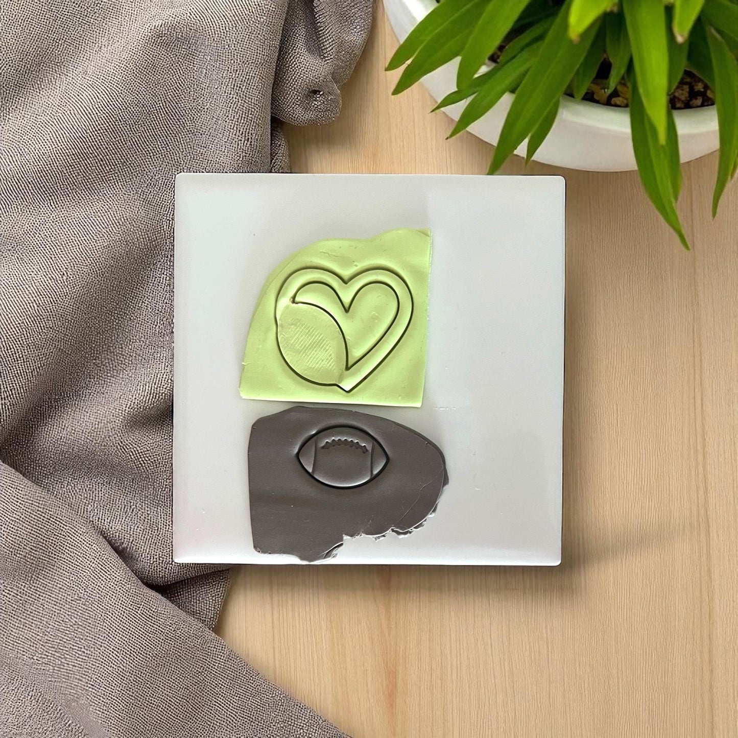 Green heart clay imprint and brown football clay imprint on white tile with fabric and plant.