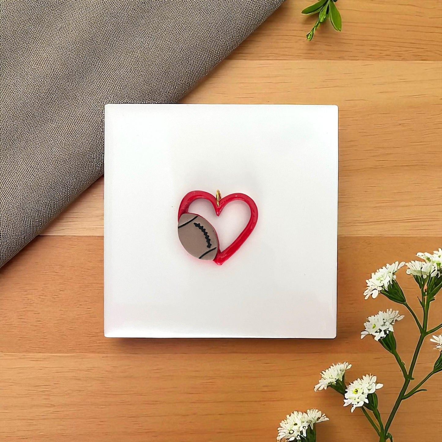 Red heart with football clay piece on white tile with fabric and flowers.