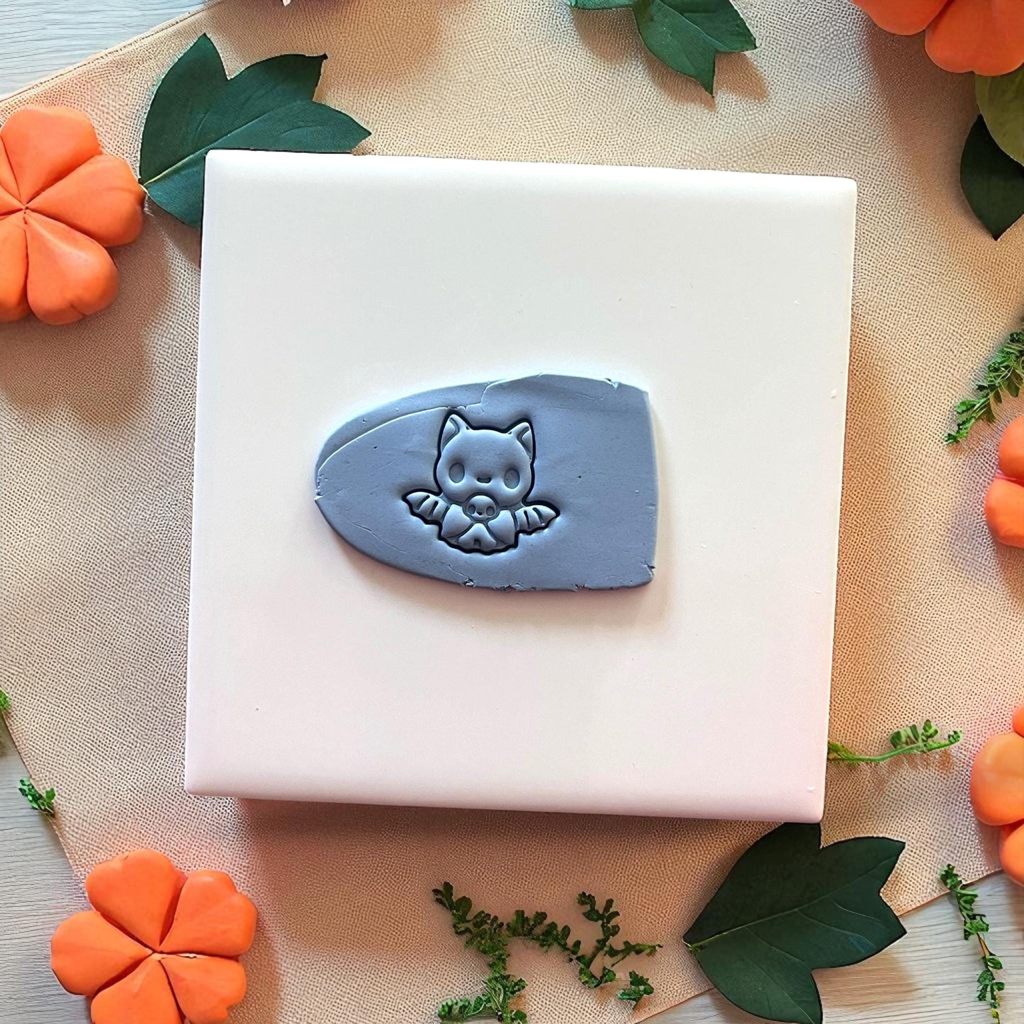 Baby bat clay imprint in gray clay on white tile with pumpkins and leaves.