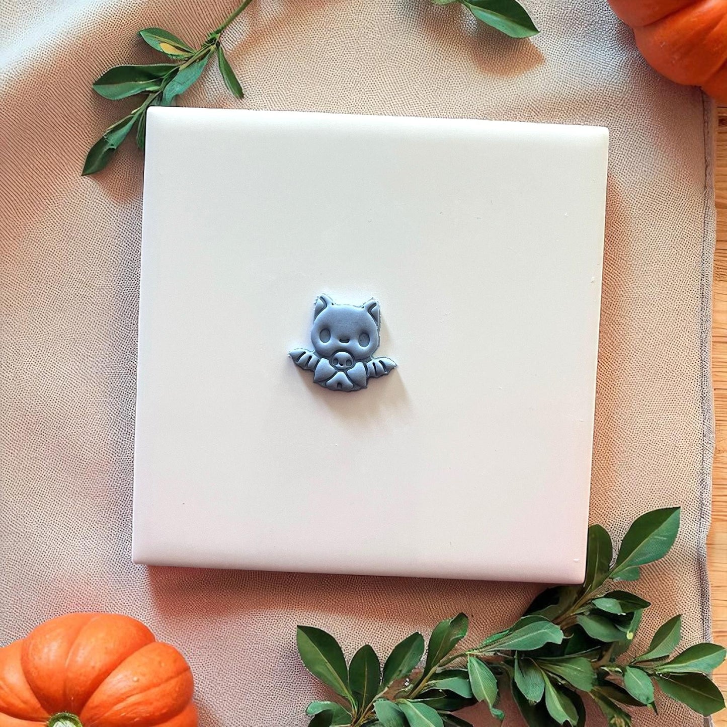 Small gray baby bat clay piece on white tile with pumpkins and greenery.