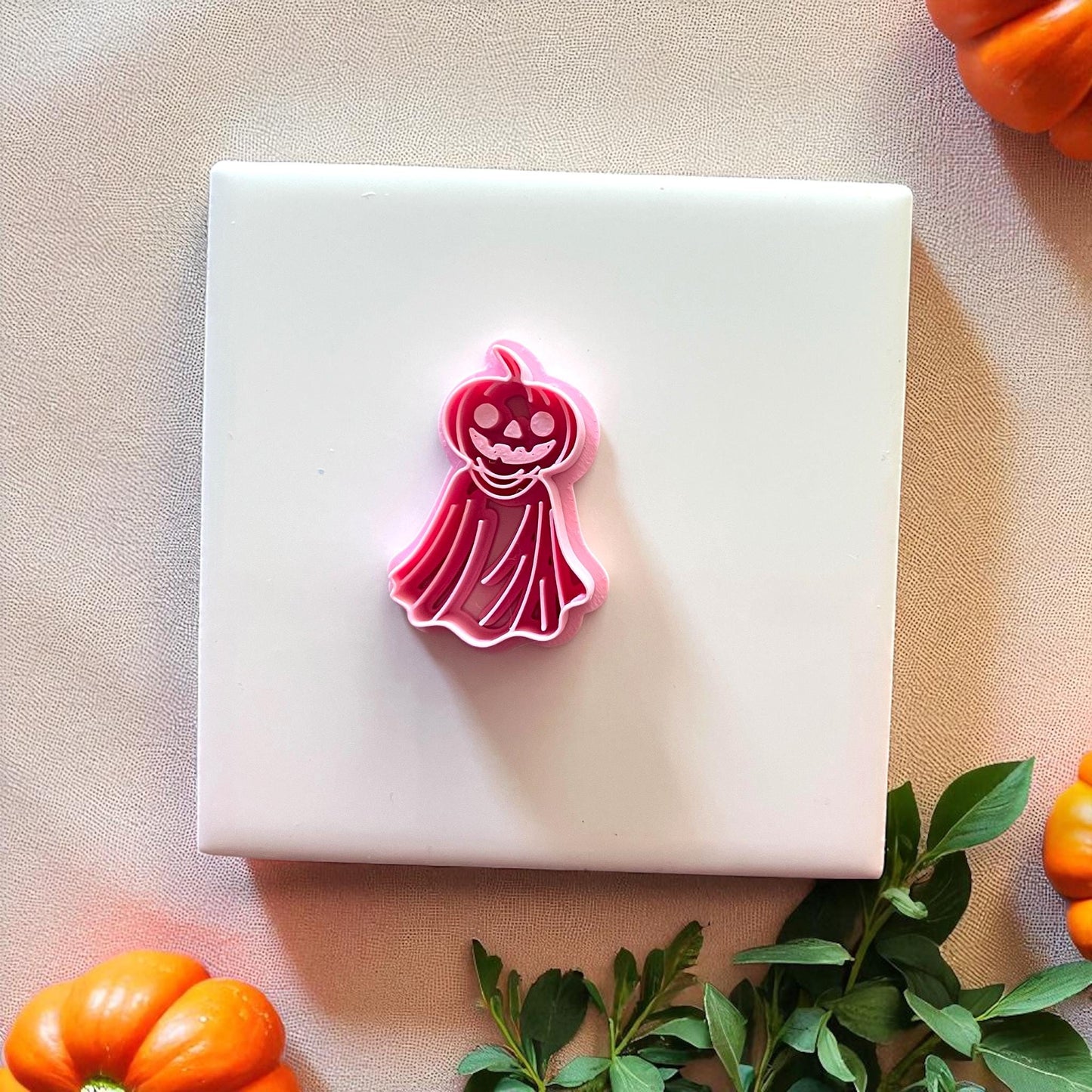 Pink pumpkin ghost cutter displayed on white tile with pumpkins and greenery.