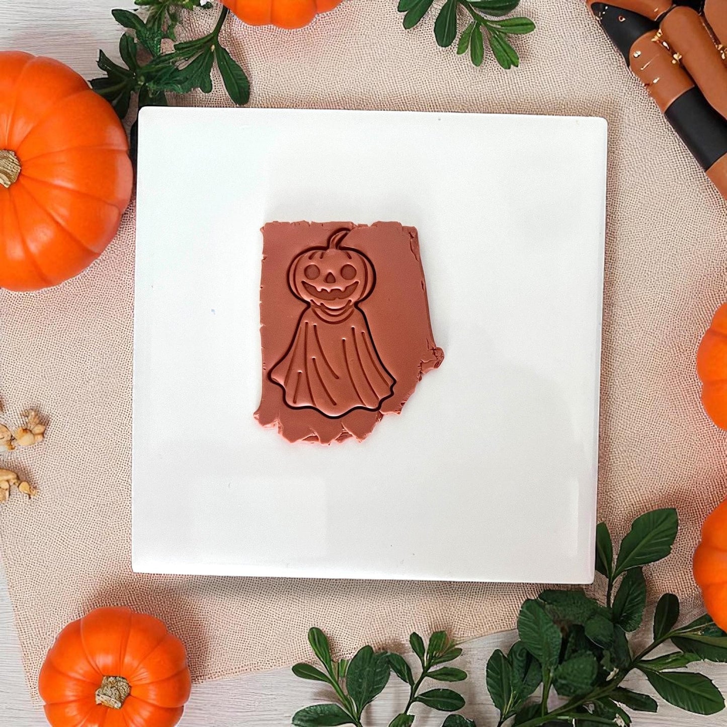 Pumpkin ghost clay imprint in orange clay on white tile with pumpkins and leaves.