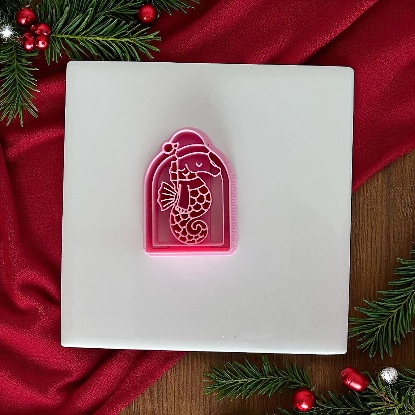 Pink Christmas seahorse clay cutter on white tile with red fabric and pine branches.