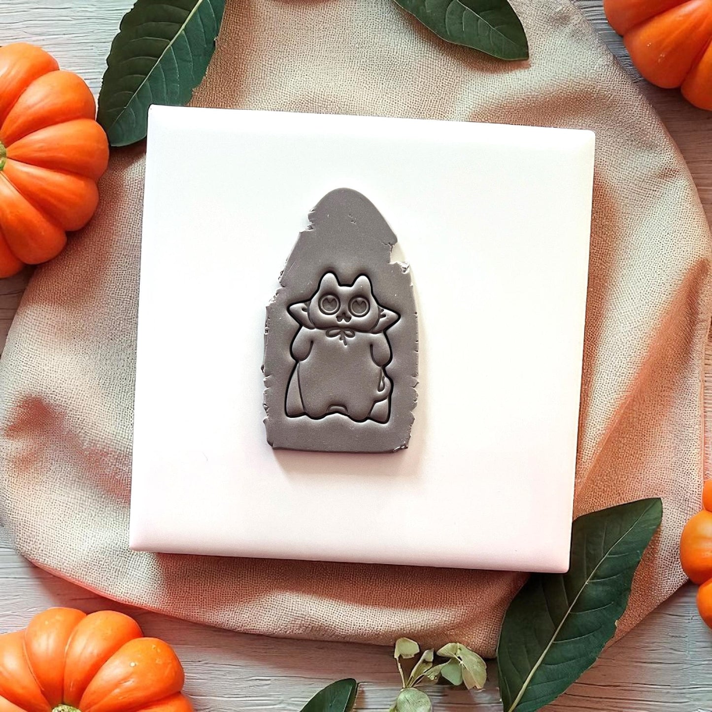 Grey Halloween cat clay imprint on white tile with pumpkins and leaves.