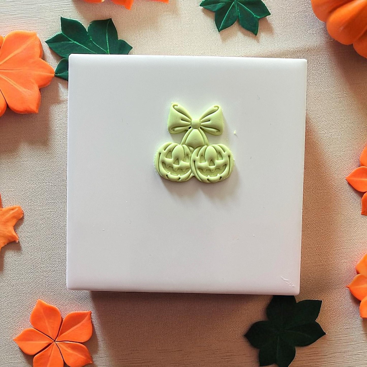Green pumpkin cherry bow clay imprint on white tile with pumpkins around.
