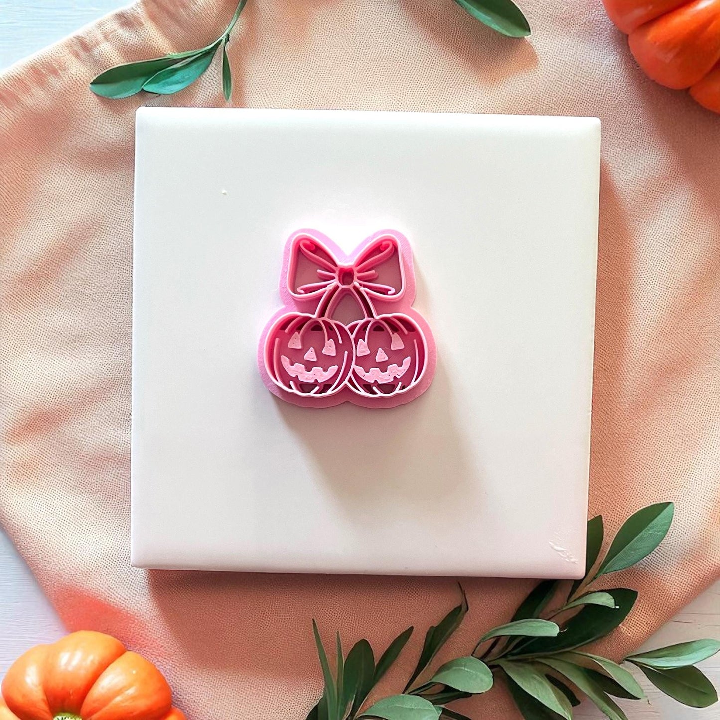 Pink pumpkin cherry bow cutter on white tile with leaves and pumpkins.