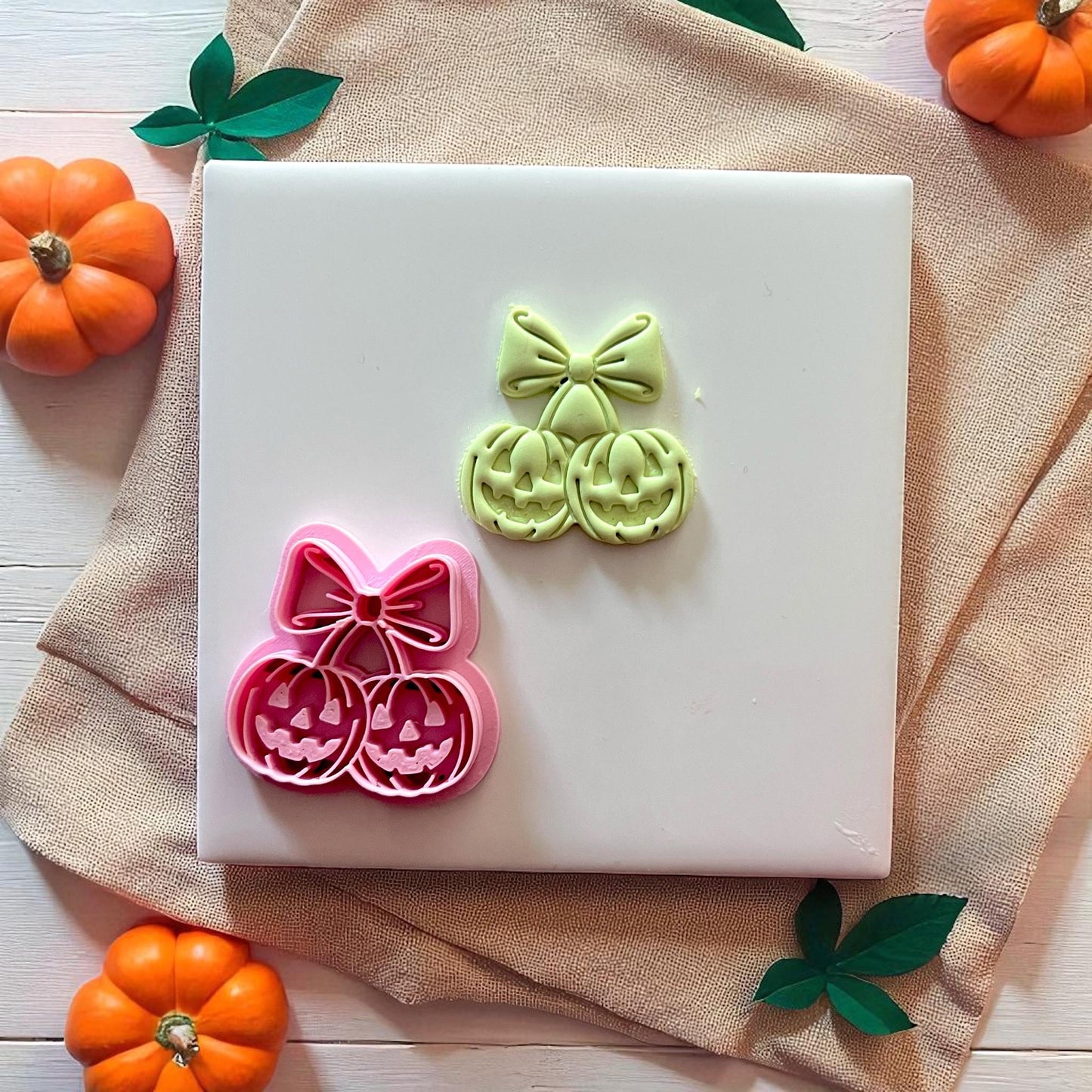 Pink pumpkin cherry bow cutter with green imprint on white tile and pumpkins.