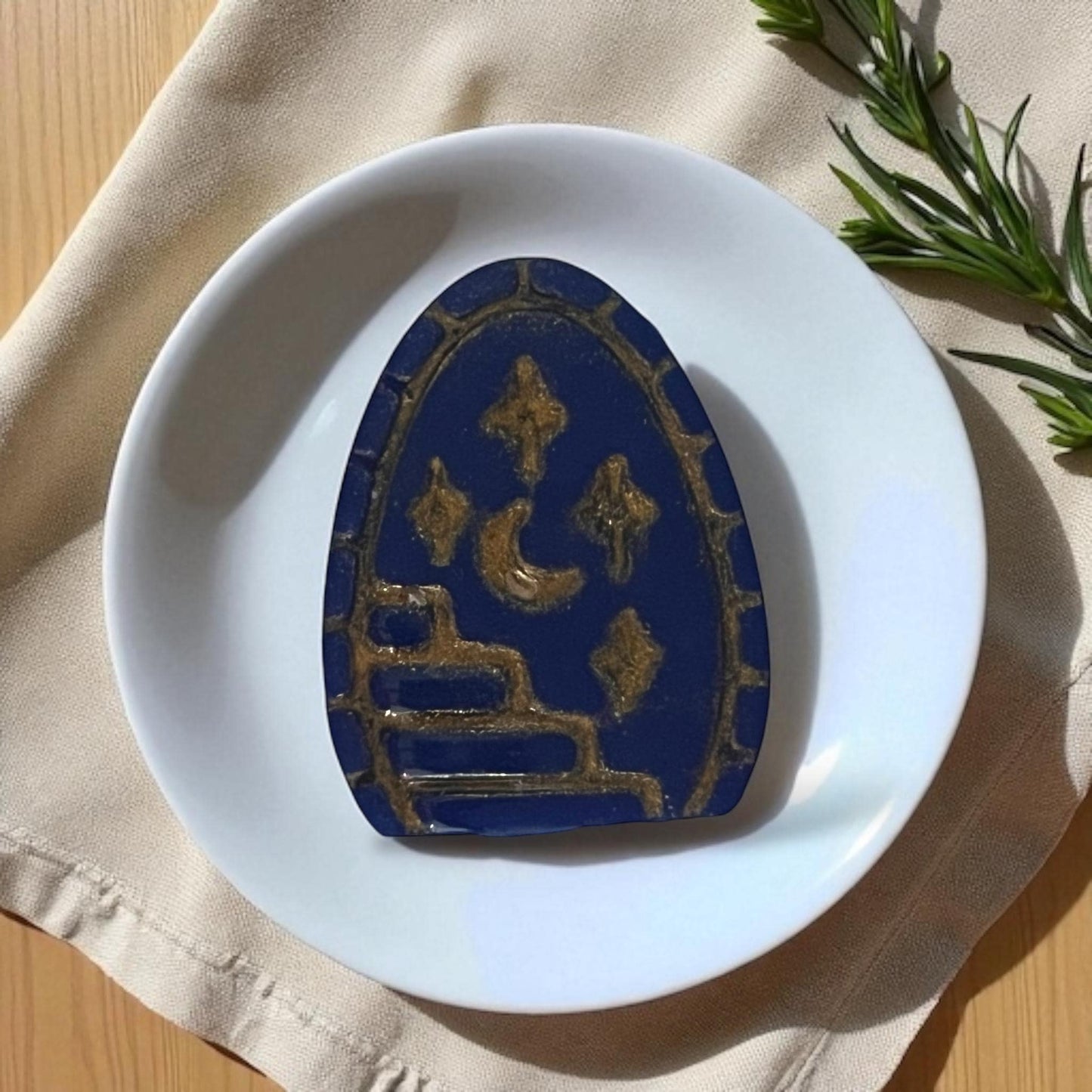 Large blue and gold arched clay piece with moon, stars, and stairway on white plate with fabric.