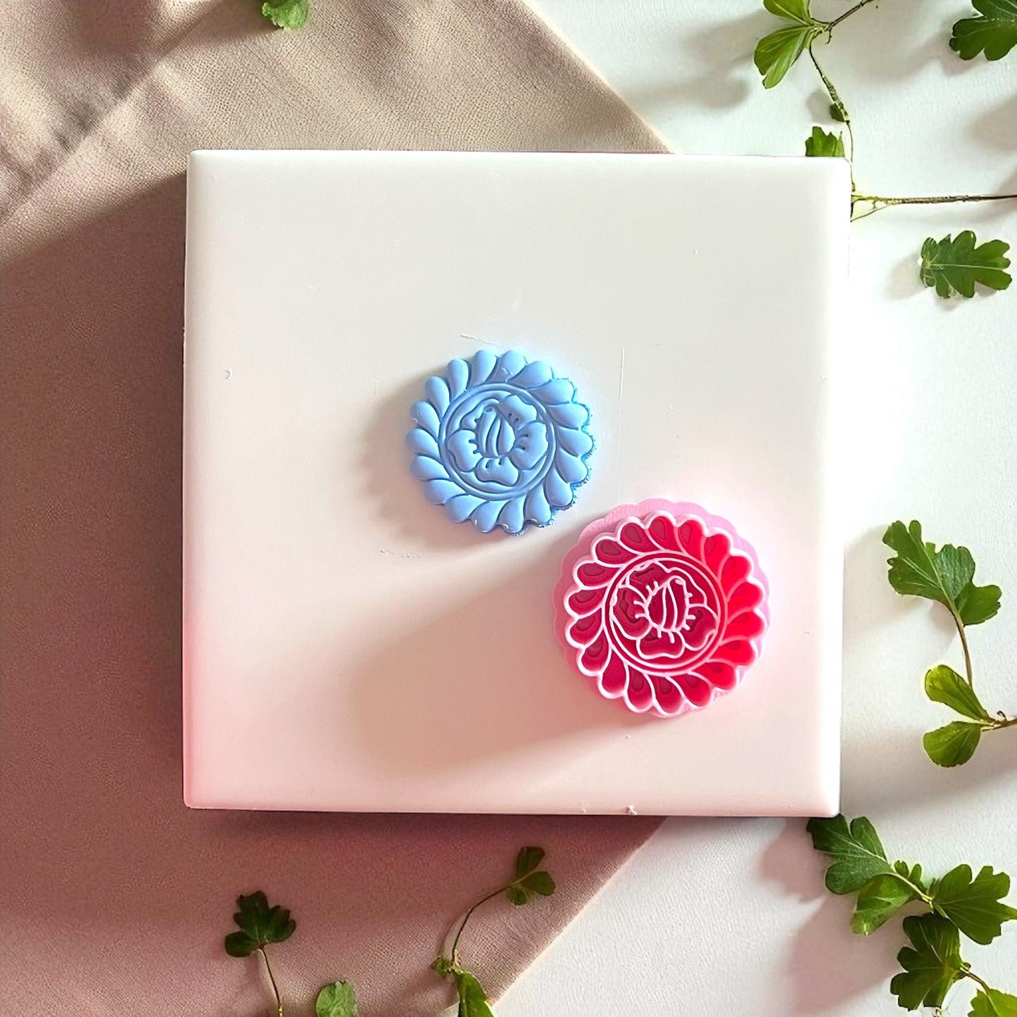 Pink flower cutter and blue clay imprint side by side on white tile with greenery.