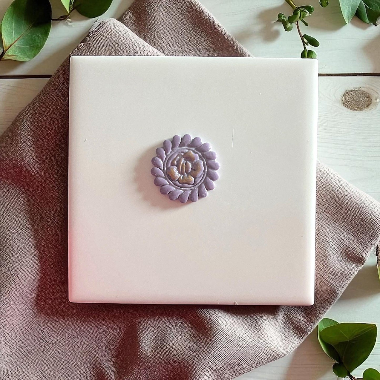 Purple clay imprint of round flower design on white tile with fabric and greenery.