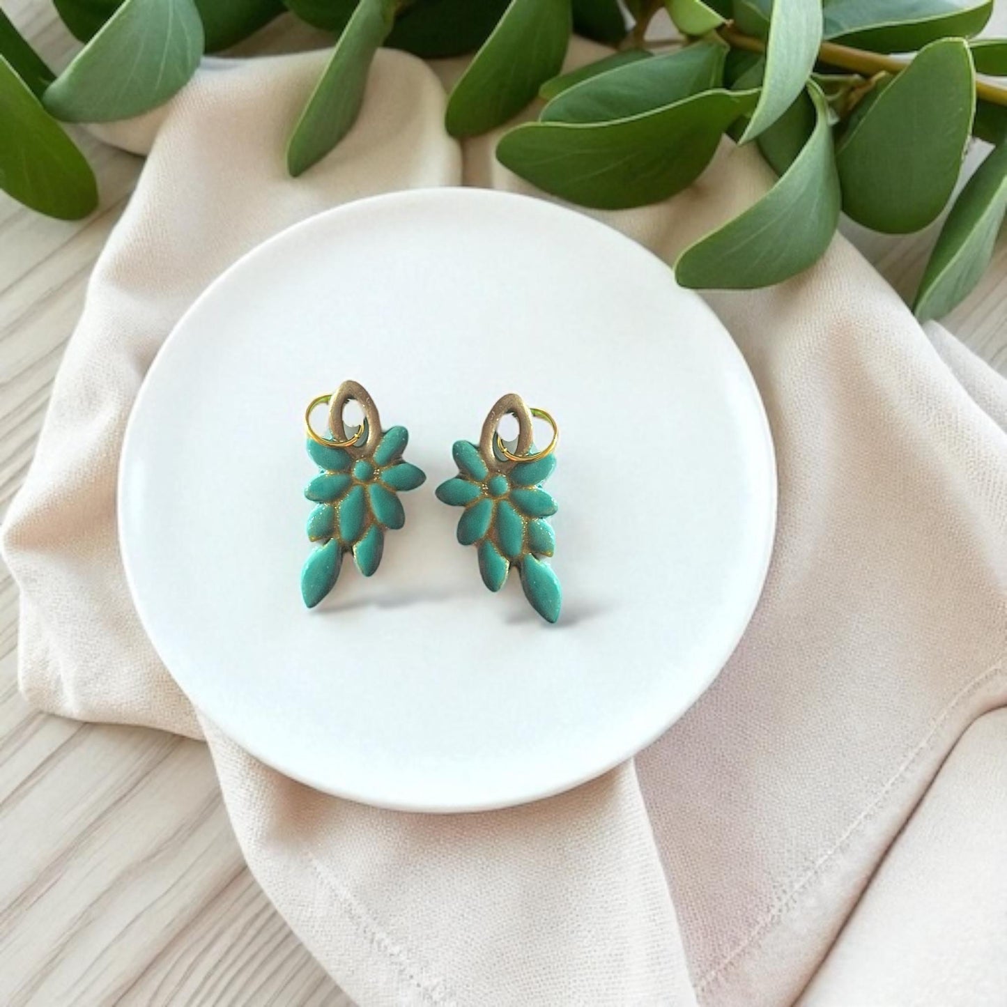 Pair of teal floral clay pieces with gold details displayed on white dish with fabric and greenery.