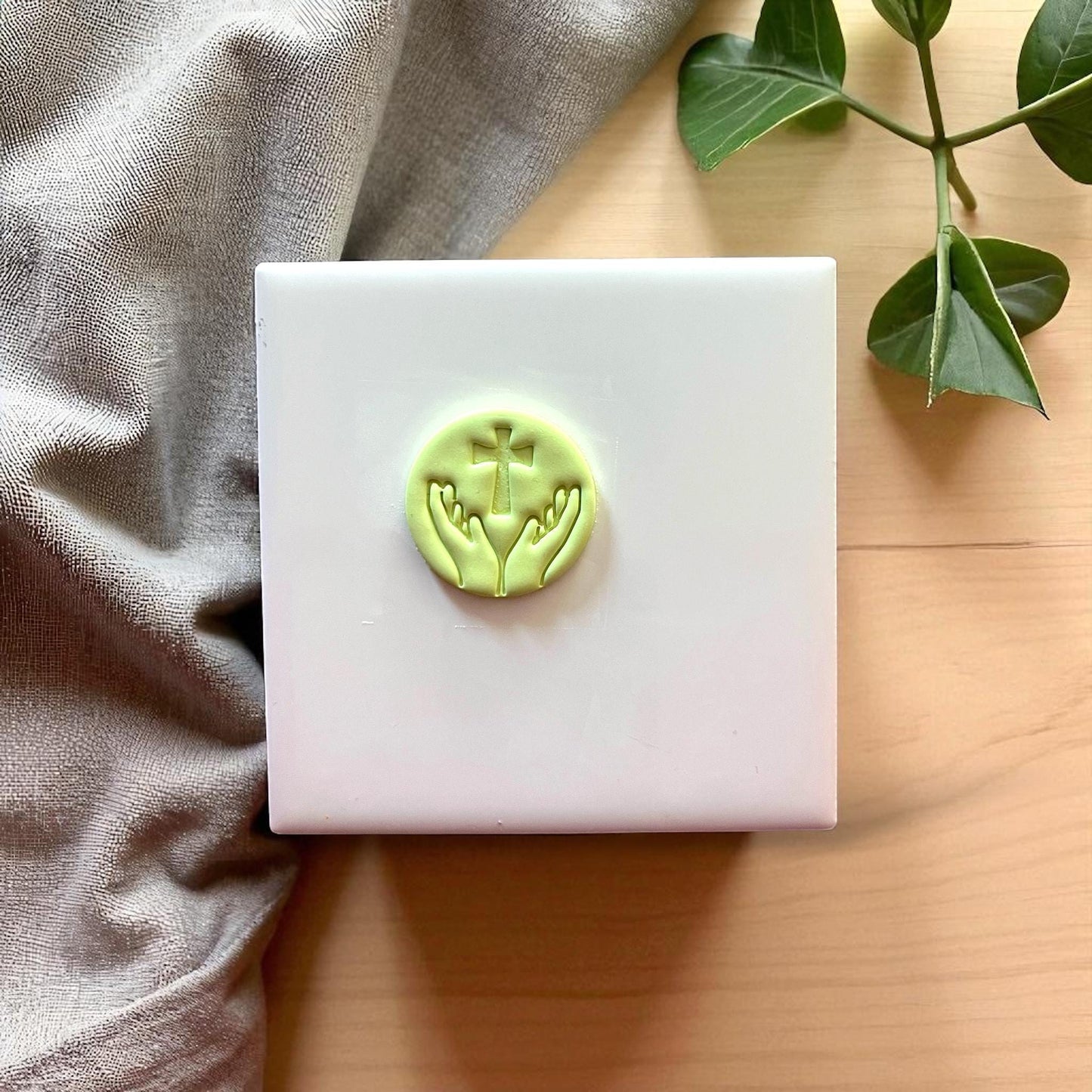 Small green clay imprint of hands lifting cross on white tile with fabric and plant.