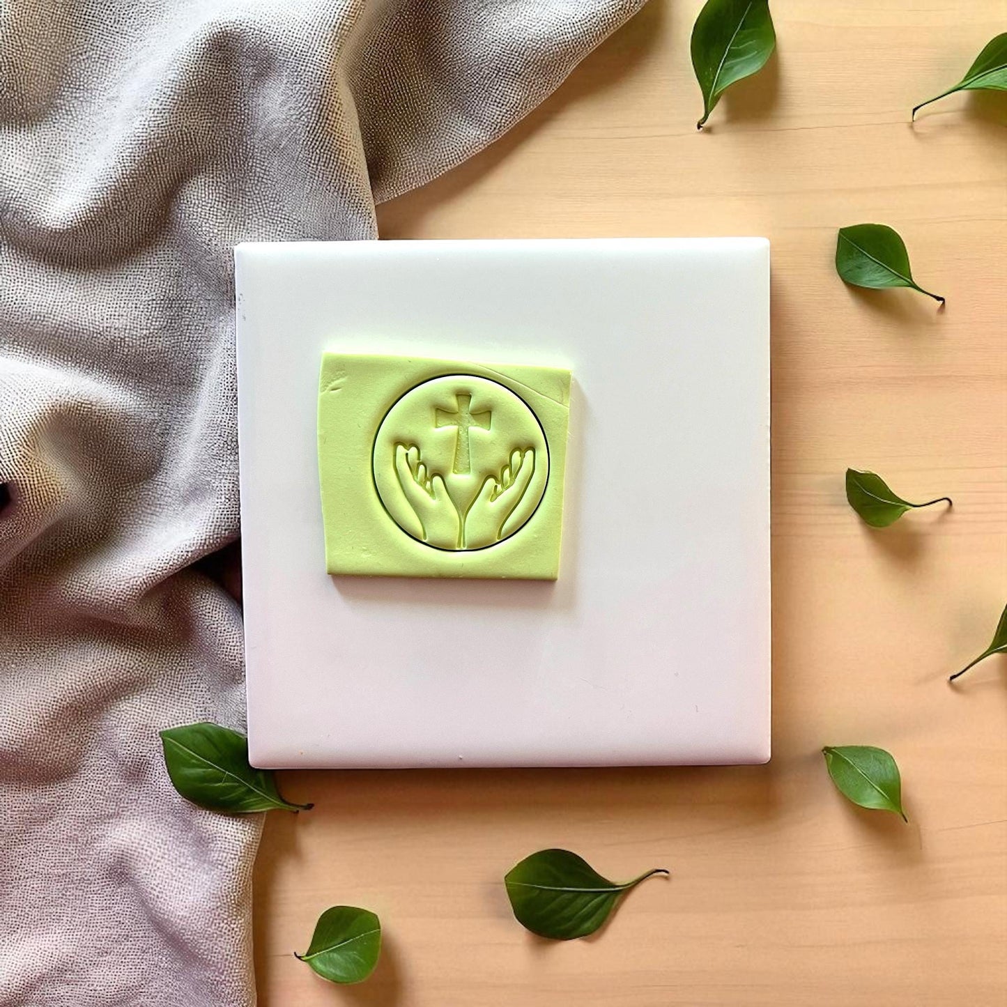 Square piece of green clay with round imprint of hands and cross on white tile with leaves.