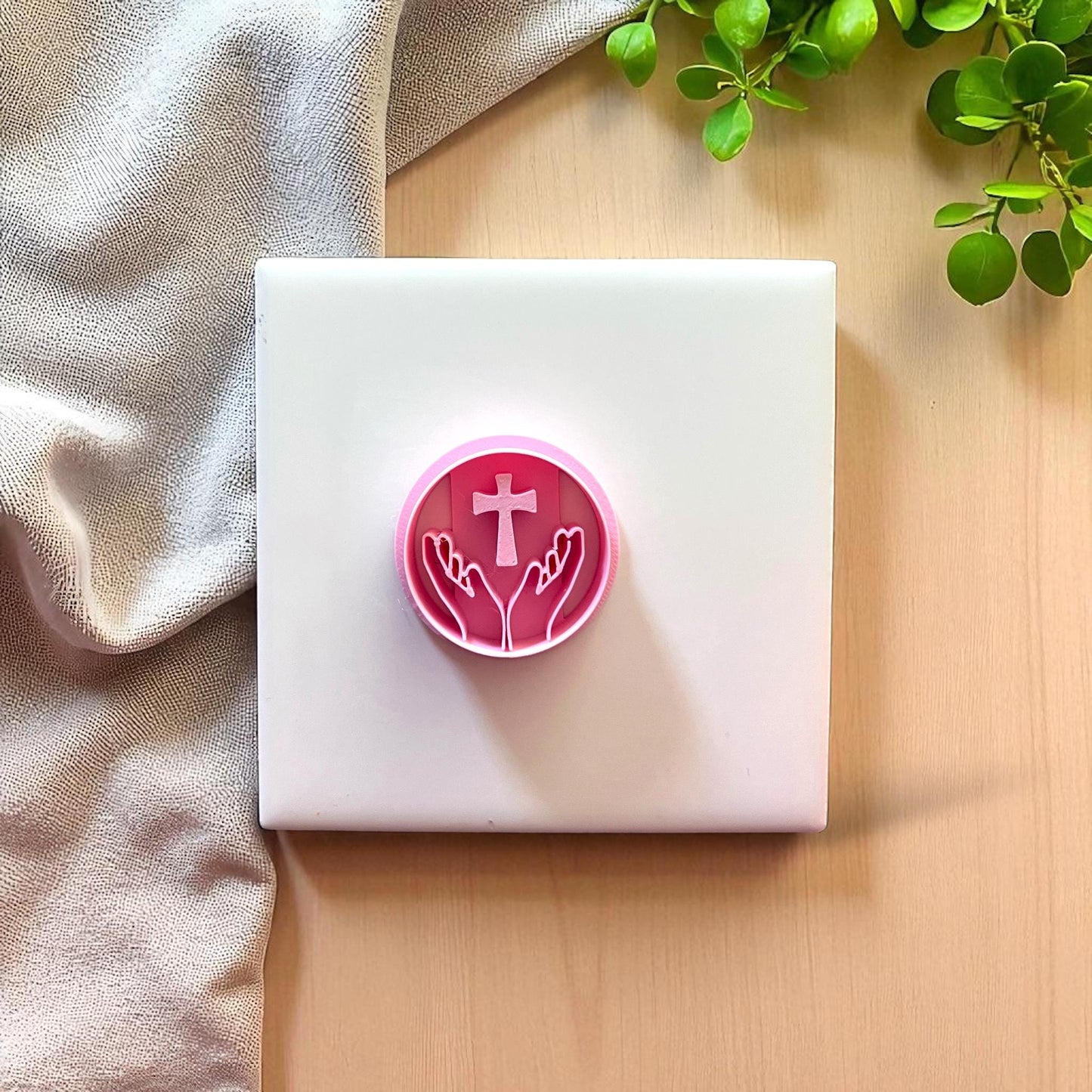 Pink round cutter with hands holding cross design on white tile with fabric and greenery.