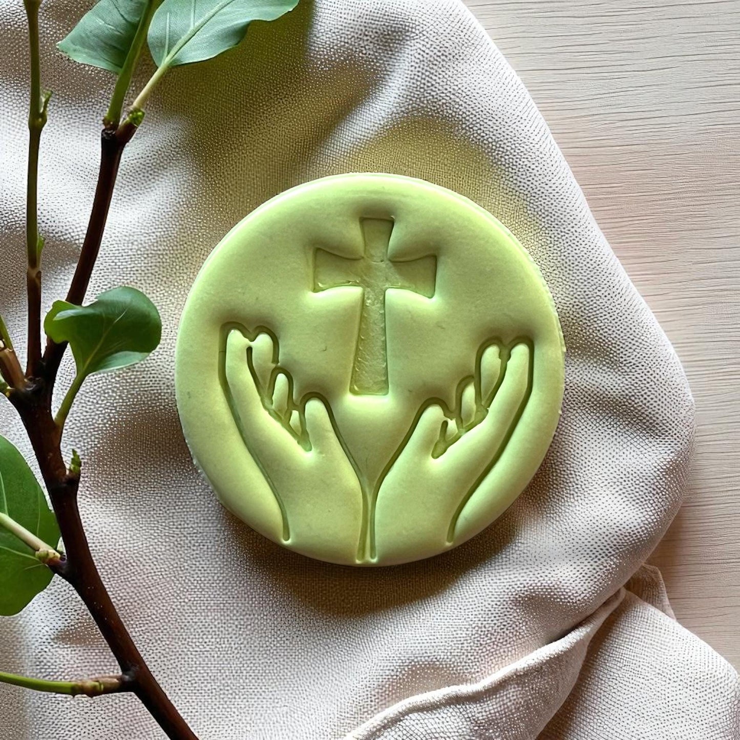 Green round clay imprint of hands lifting cross on fabric with branch detail.
