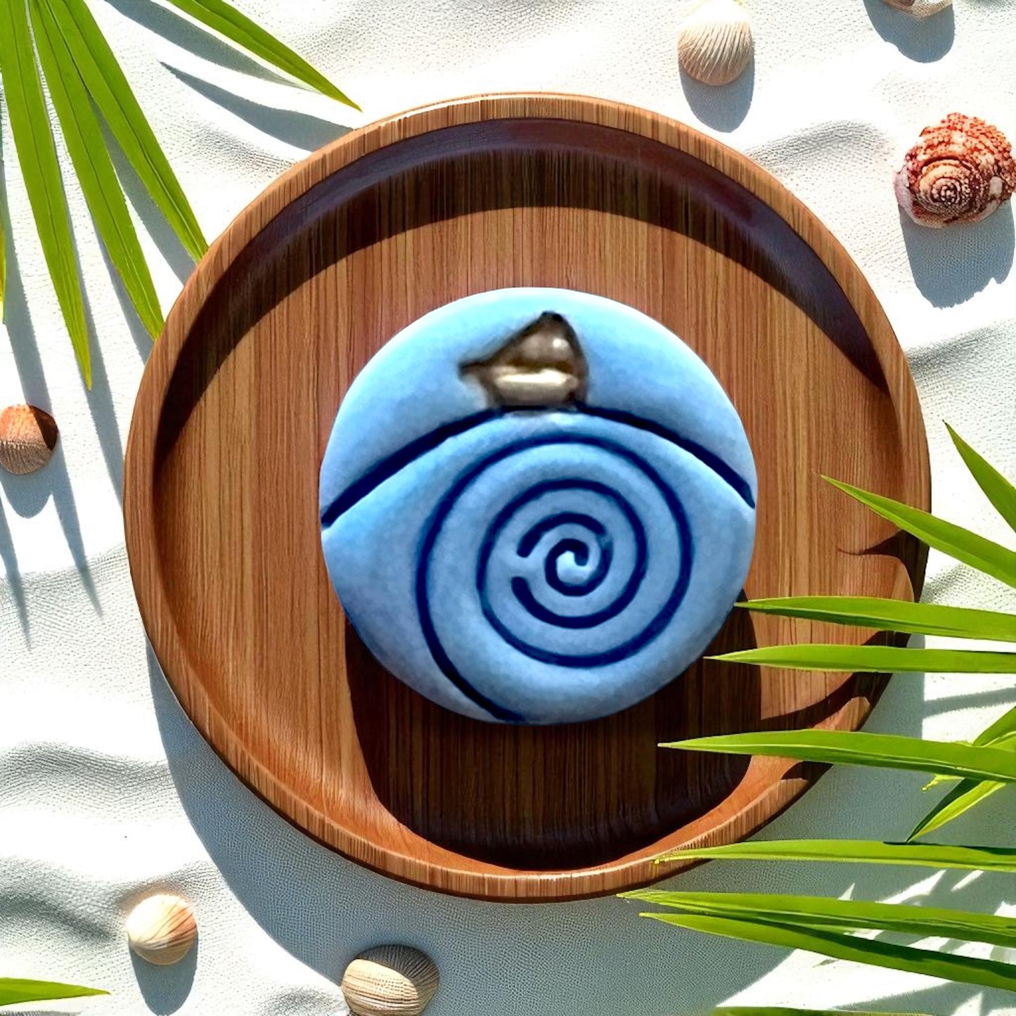 Blue clay piece with spiral wave and paper boat detail on wood dish, shells and palm leaves.
