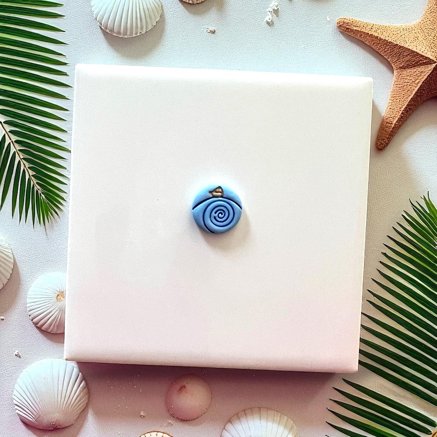 Small blue paper boat in waves clay piece on white tile with seashells, palm leaves, and starfish.