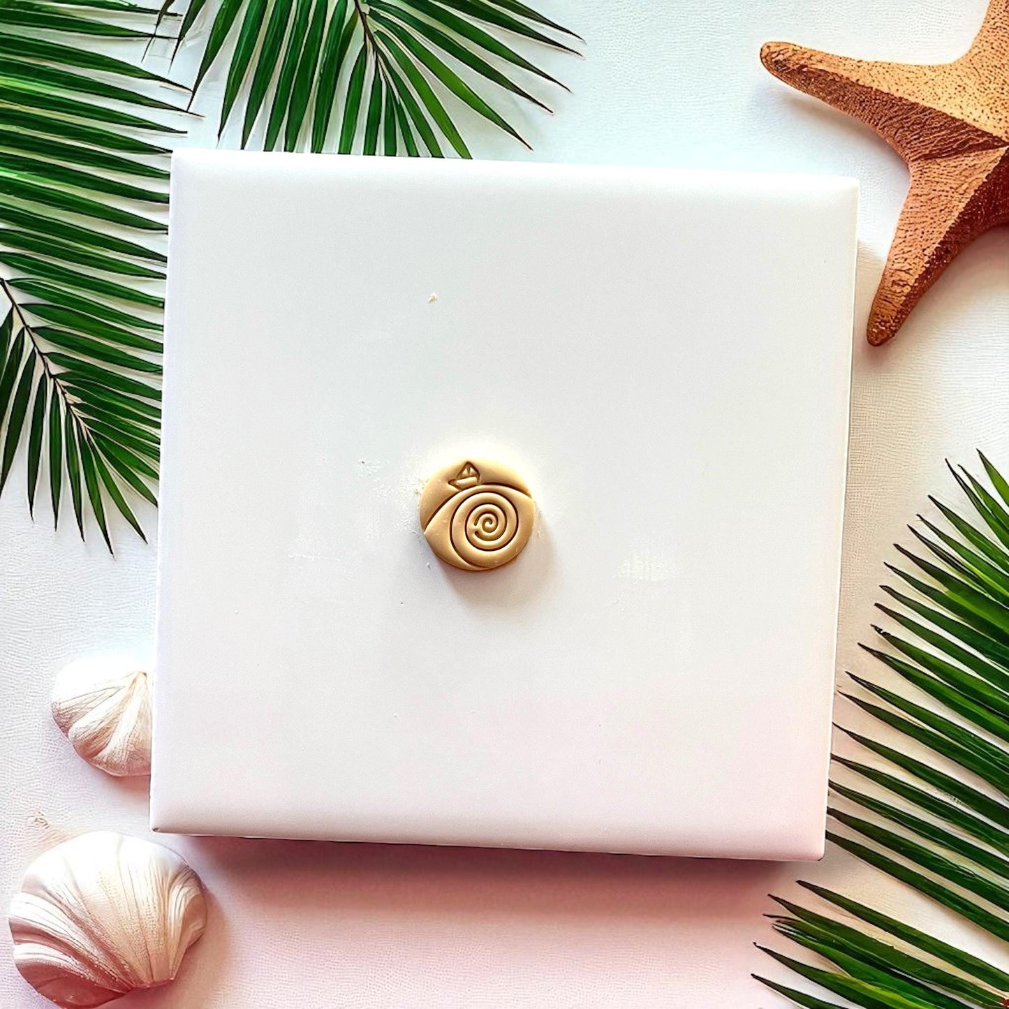 Tan clay piece showing paper boat riding spiral waves on white tile with palm leaves.