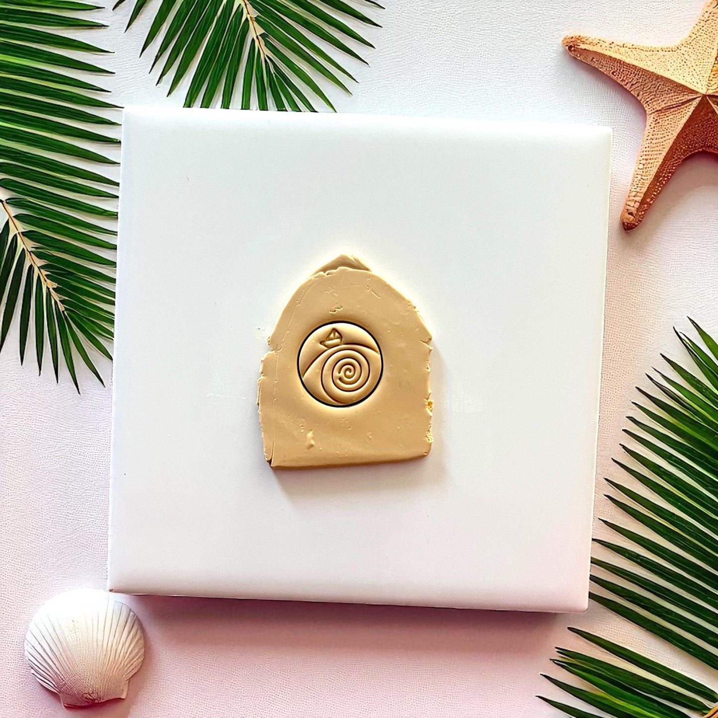 Tan clay imprint of paper boat in waves design on white tile with palm leaves and starfish.