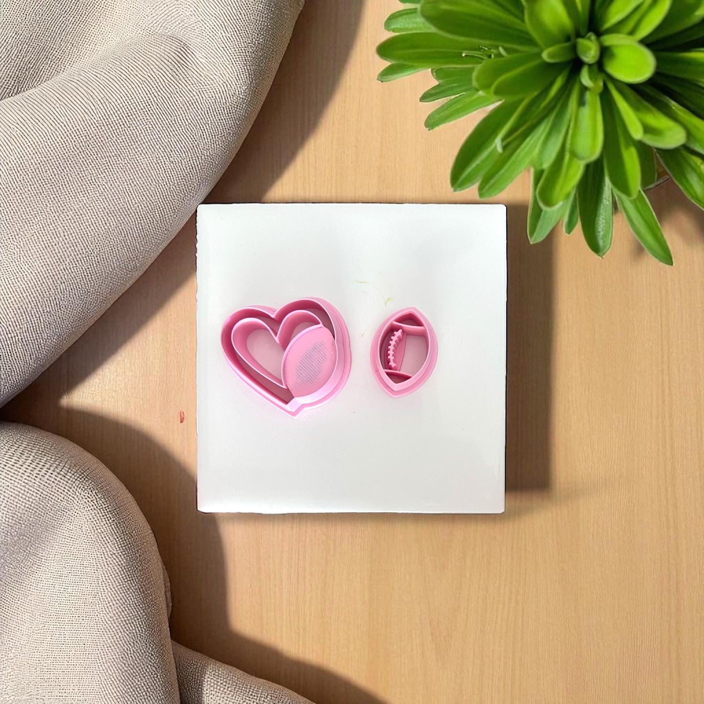 Pink football heart cutter set on white tile with fabric and green plant.