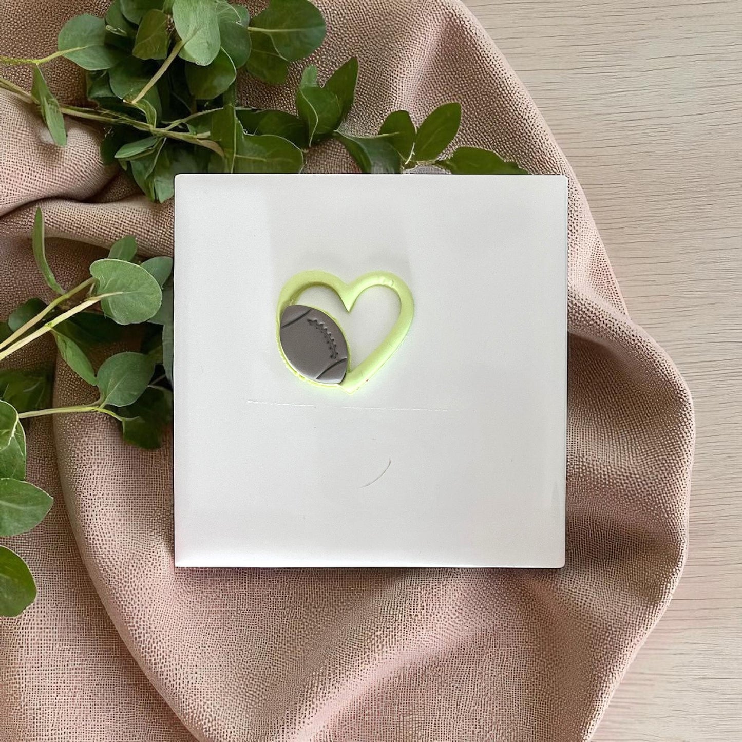 Green football heart clay piece on white tile with fabric and leafy background.