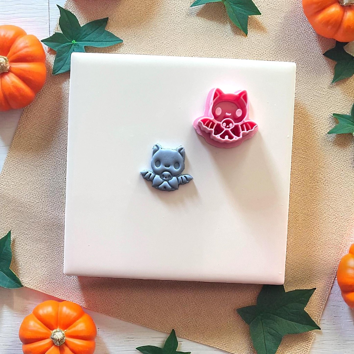 Gray baby bat clay piece beside pink bat cutter on white tile with pumpkins.