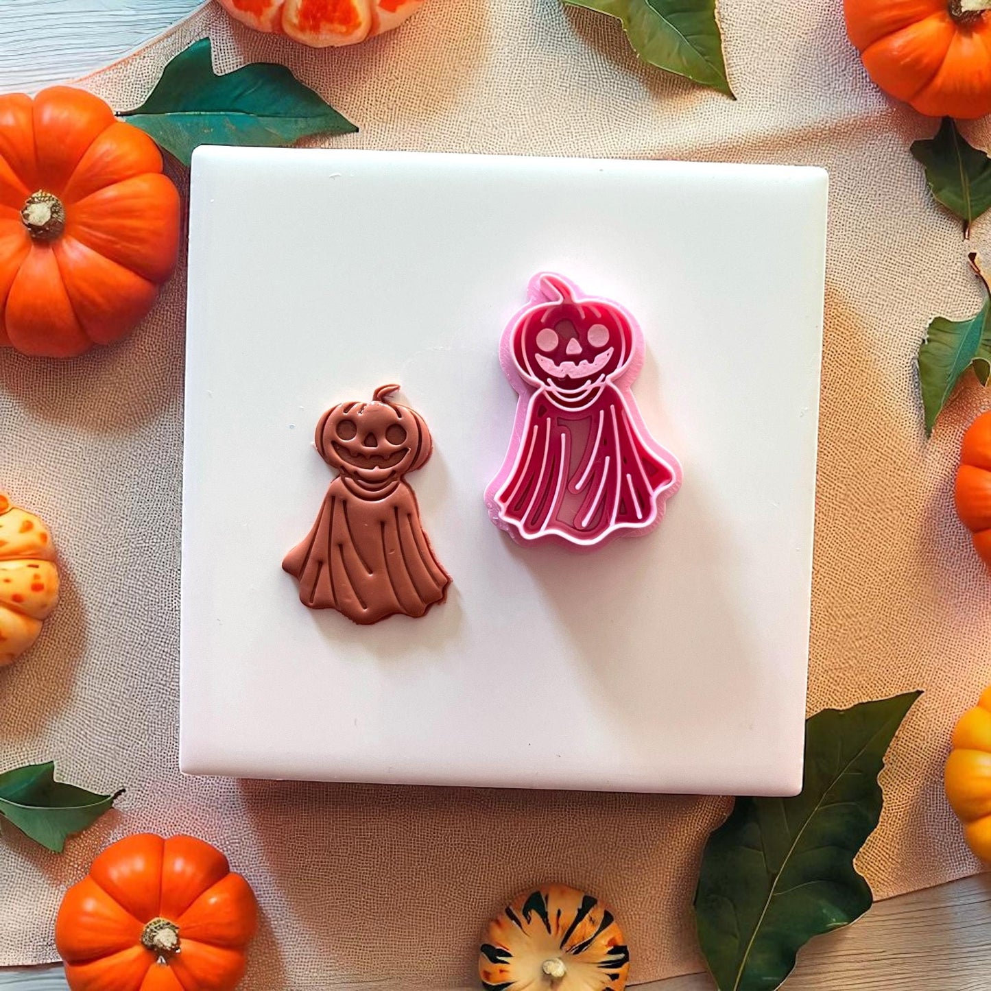 Orange pumpkin ghost clay piece beside pink cutter on white tile with pumpkins.