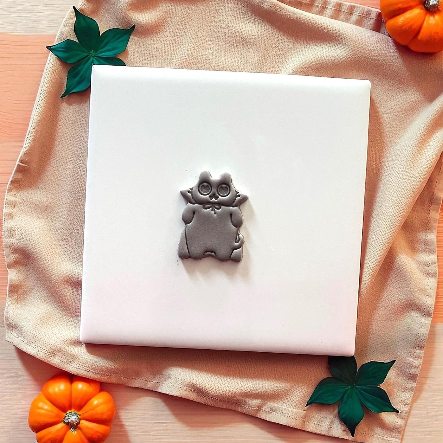 Grey cat clay imprint on white tile with beige cloth and pumpkins.