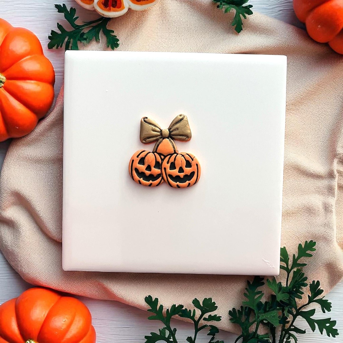 Orange pumpkin cherry clay imprint with gold bow on white tile and pumpkins.