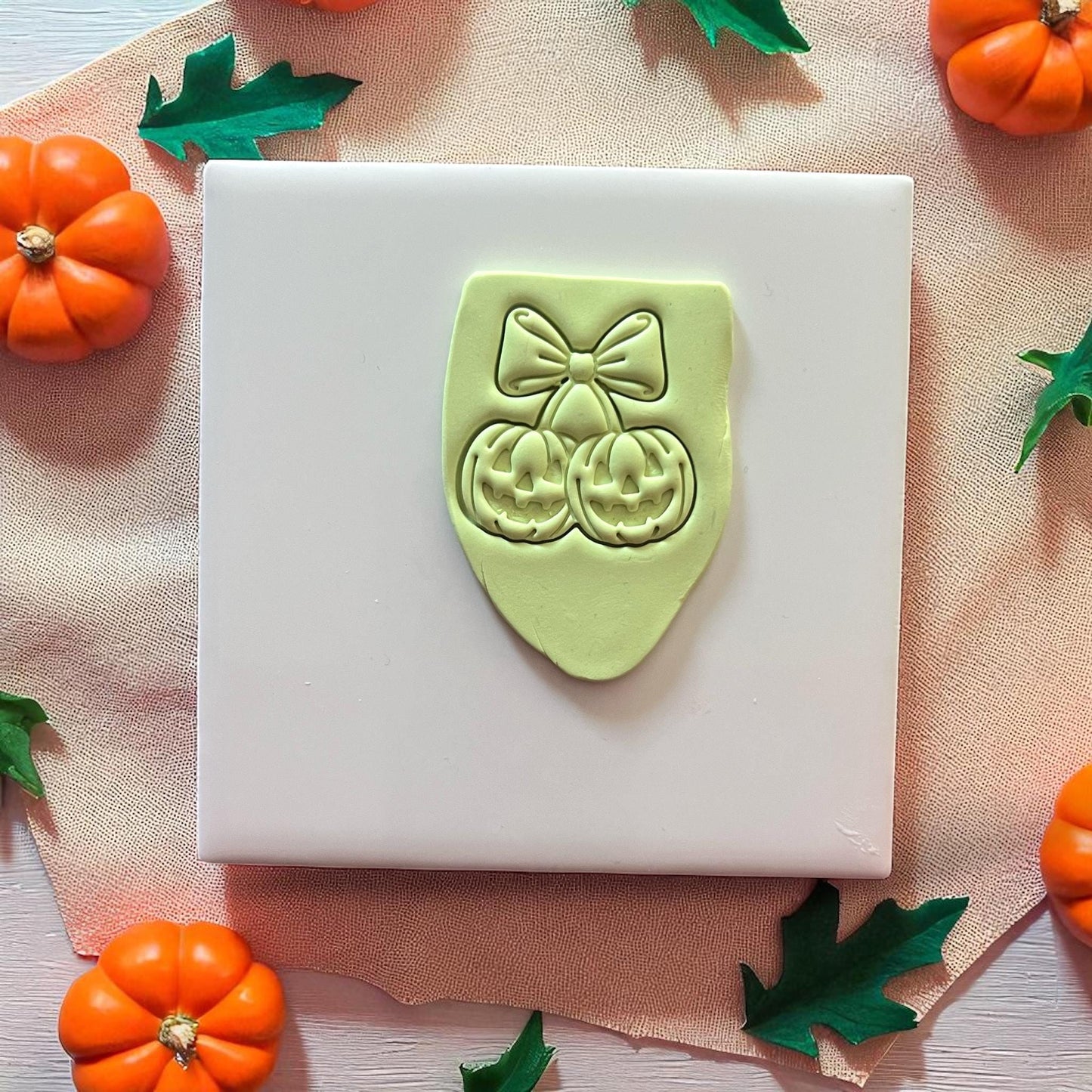 Green pumpkin cherry bow clay imprint on white tile with orange and green leaves.
