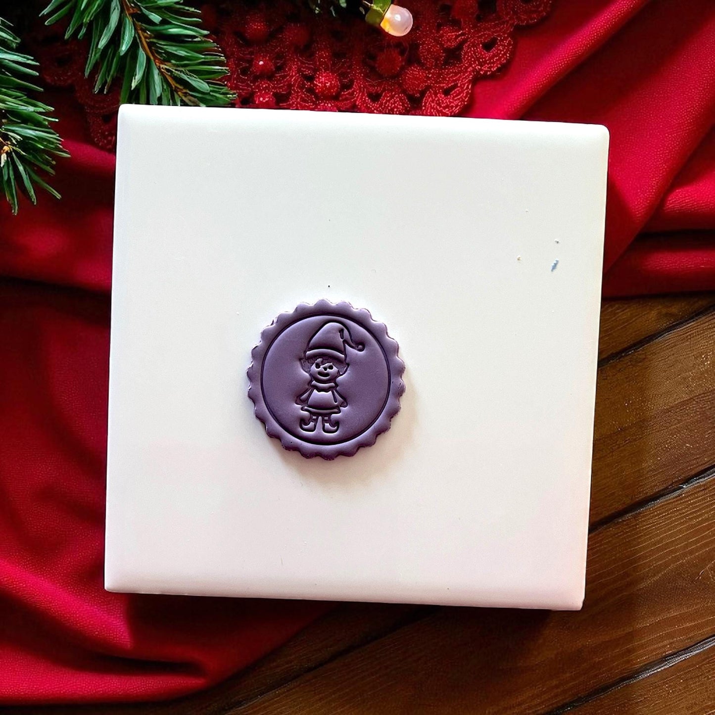 Small purple elf clay imprint on white tile with red cloth and pine.