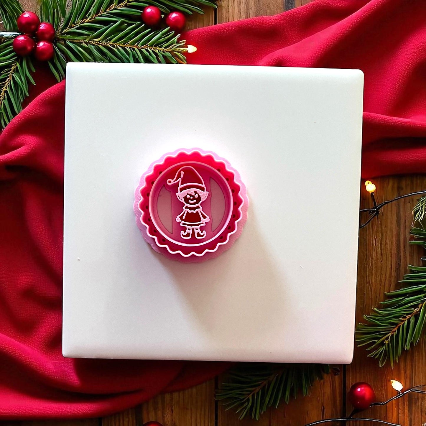 Pink scalloped circle elf clay cutter on white tile with red fabric and pine branches.