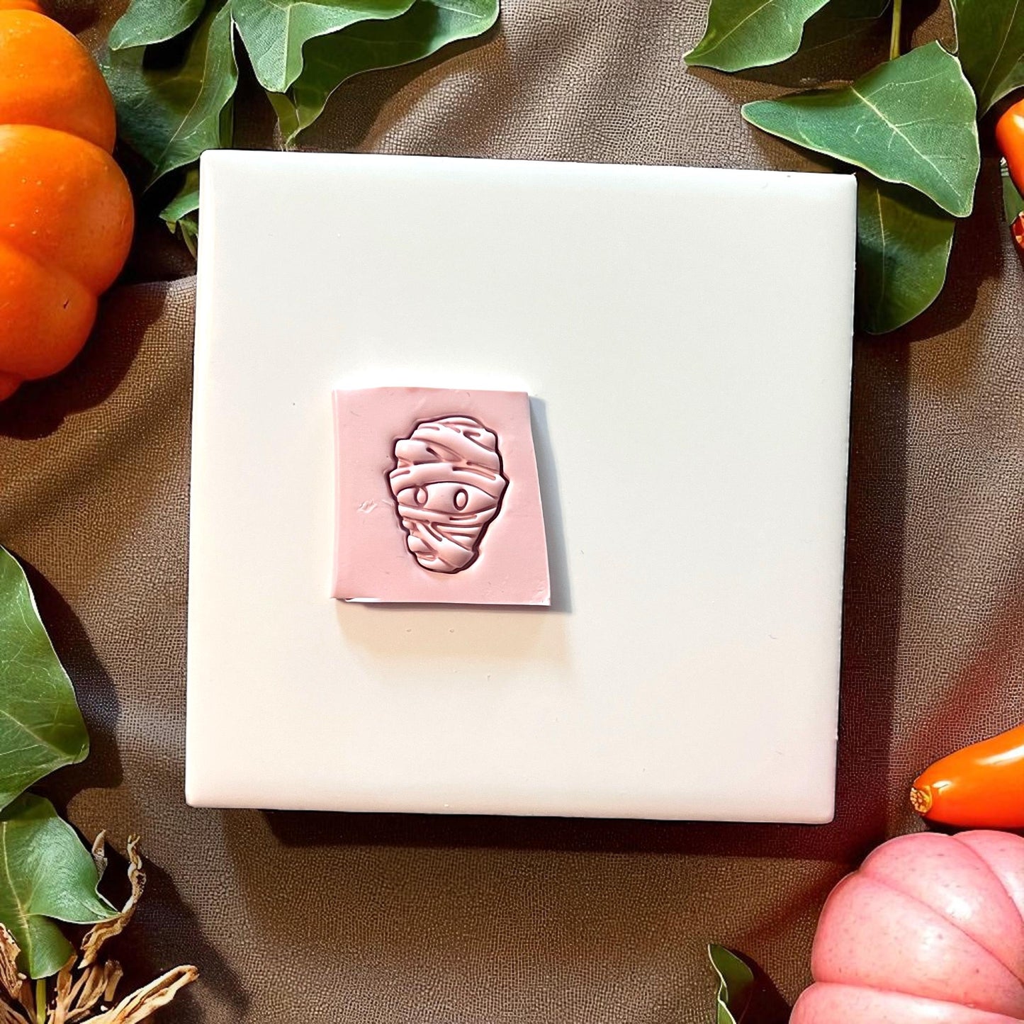 Pink mummy clay impression on square slab with pumpkins and greenery
