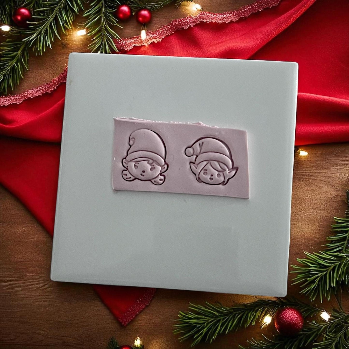 Polymer clay imprints of elf boy and girl faces on white tile with red holiday fabric