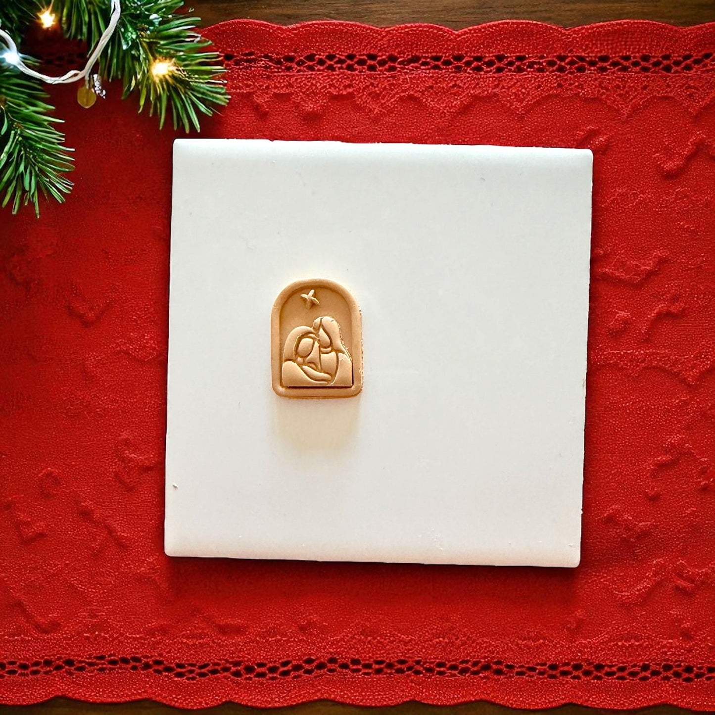 Small Nativity clay piece centered on white tile with red lace runner and pine