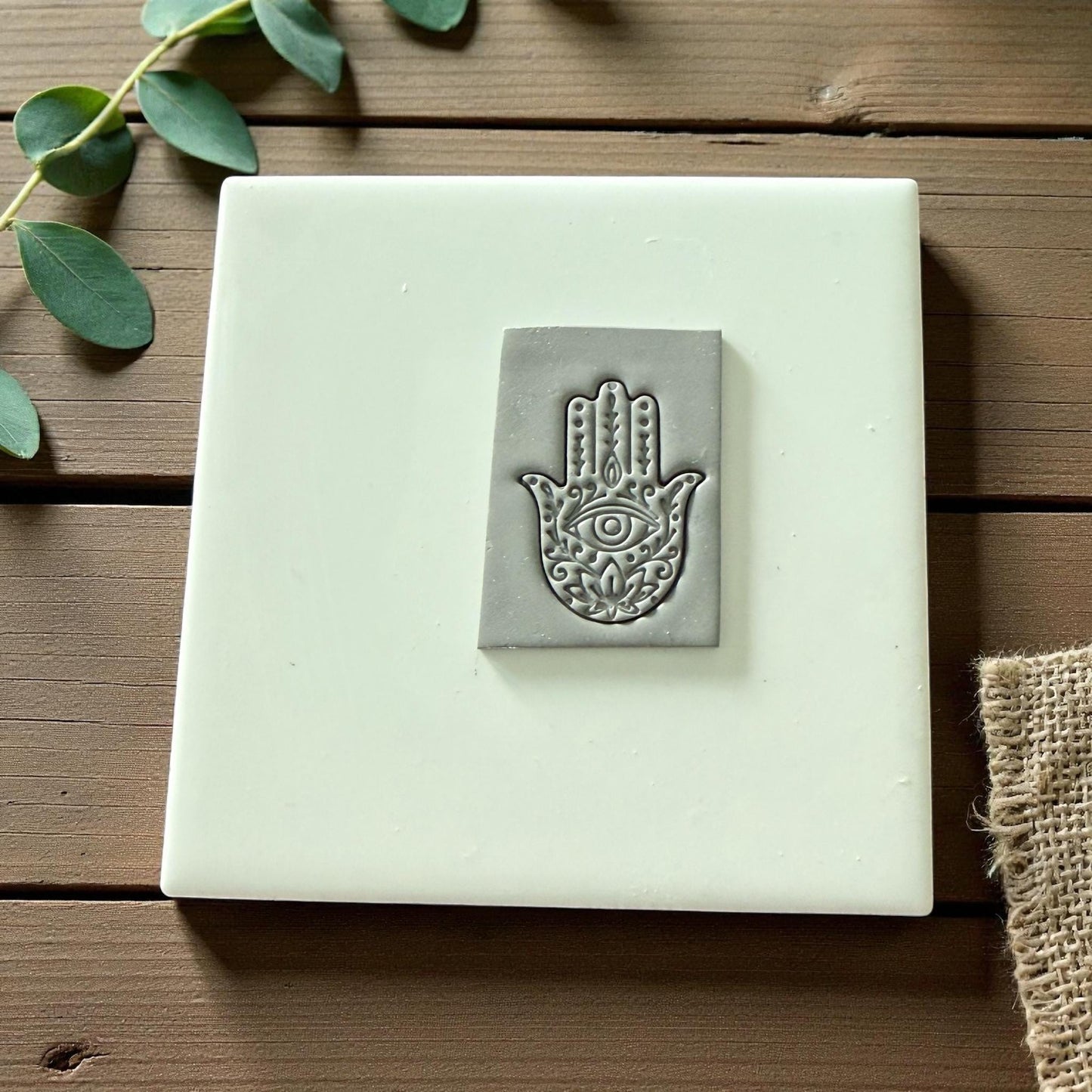 Gray Hamsa imprint pressed into clay sheet on white tile with beige fabric