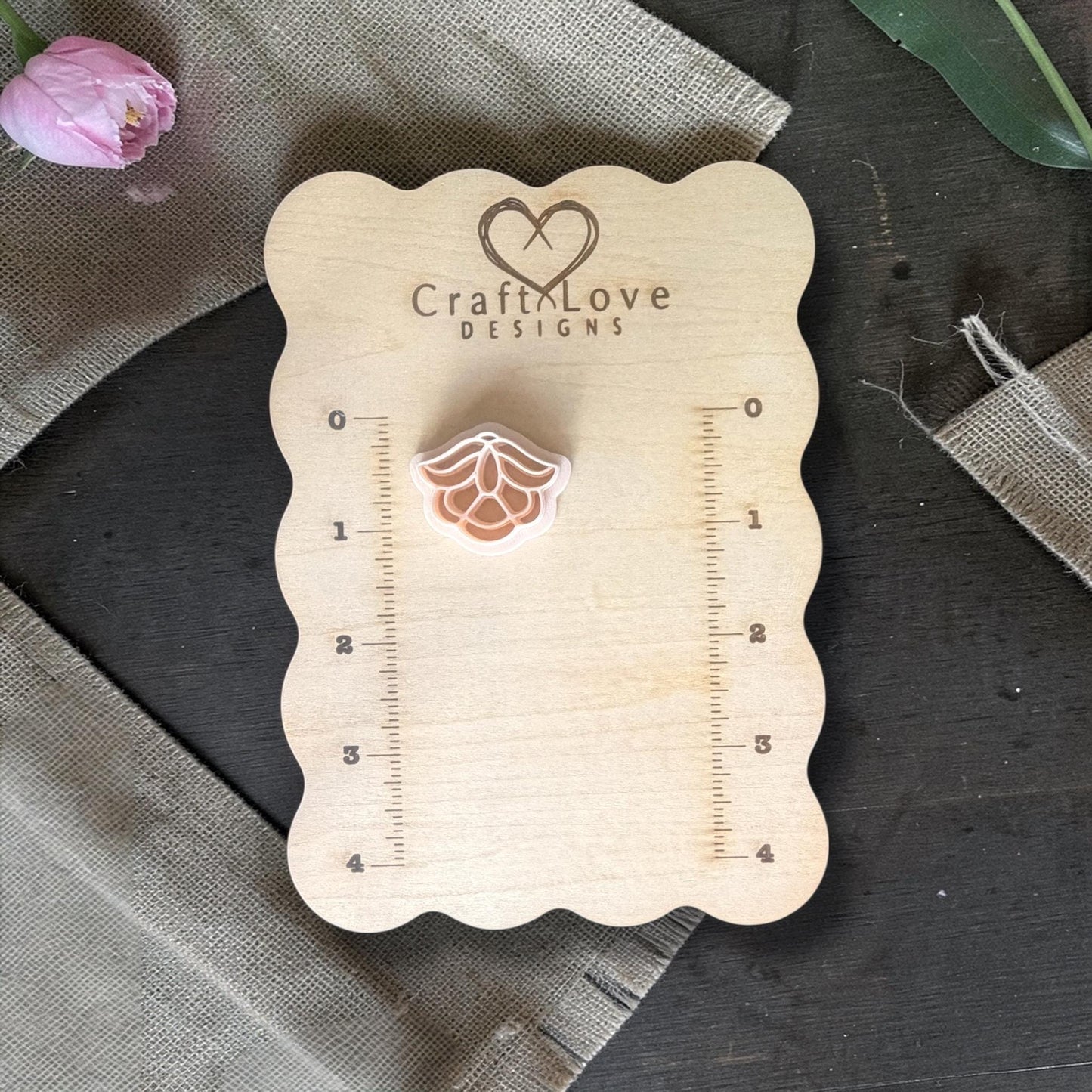 Small floral polymer clay cutter placed on a wooden CraftLove Designs measuring board with burlap fabric and a pink flower beside it.